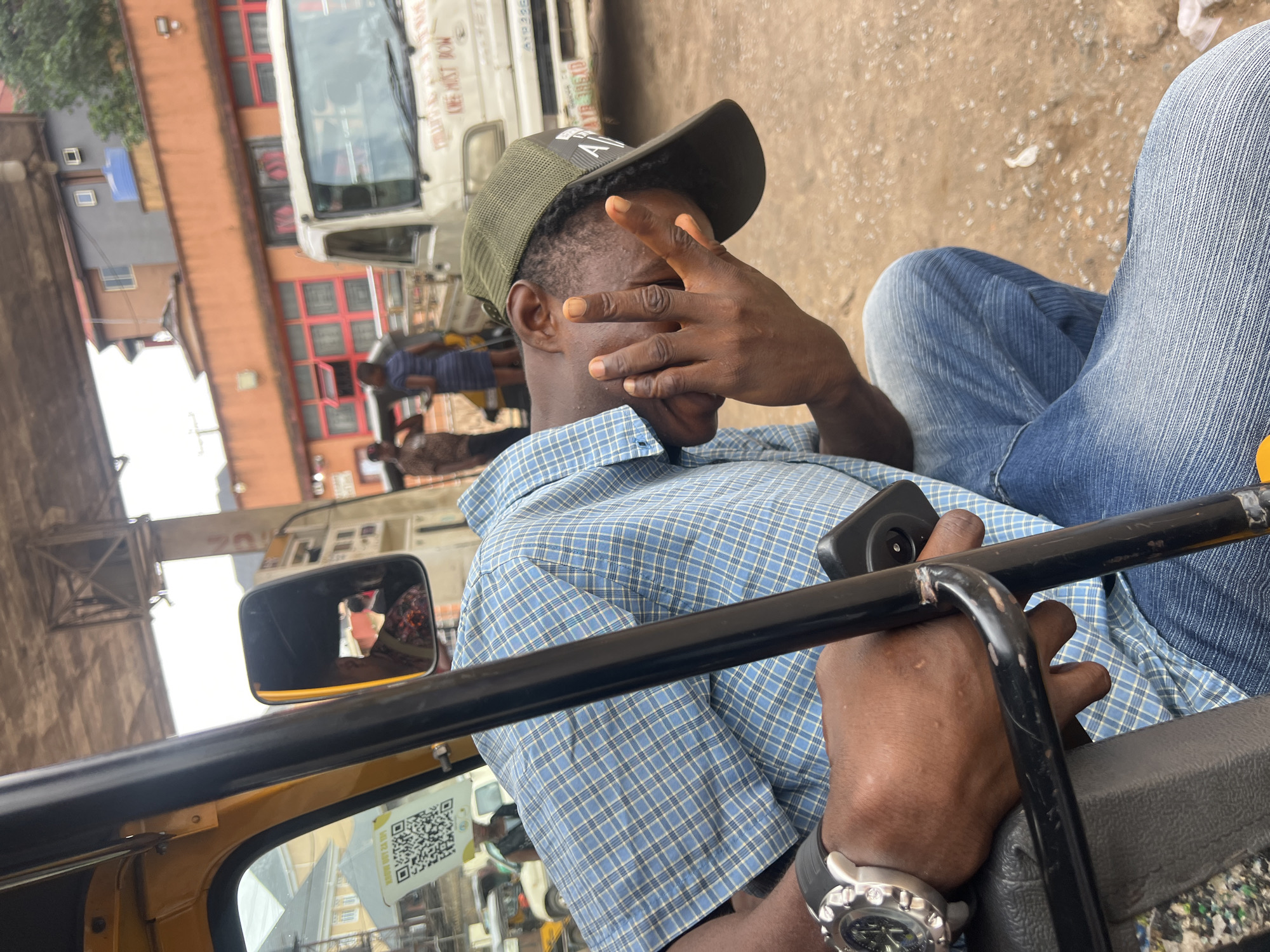 A medium shot captures a Black man sitting in the driver's seat of a yellow auto-rickshaw in Nkpor, Nigeria. He is wearing a blue and white checkered shirt, a green baseball cap, and blue jeans. His right hand is raised, covering his face as if shielding his eyes from the sun or posing for the photo. A black smartphone is held in his left hand, resting on his lap. A silver watch with a black band is visible on his left wrist.
The background depicts a bustling street scene with buildings and vehicles. To the left, a white truck with red accents and text is partially visible. A building with orange and red facade and multiple windows is also present. The ground is sandy and appears dry, suggesting a sunny day. A side-view mirror of the auto-rickshaw is in the foreground, reflecting the surroundings, including a glimpse of the driver's face and the scene behind. A QR code sticker is affixed to the windshield. The overall impression is that of a typical day in a busy Nigerian urban environment. The man's gesture of covering his face could suggest a desire for privacy or a playful interaction with the photographer.