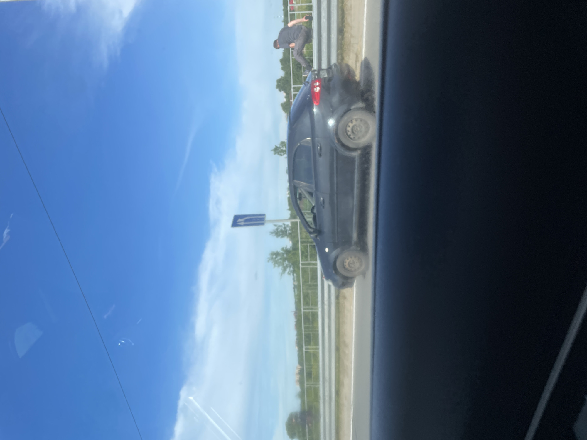 The image shows a dark-colored sedan driving on a paved road under a bright blue sky with scattered clouds. A person is seen sitting on a metal guardrail next to the road, with their legs extended. The person is wearing a dark t-shirt and light-colored pants. The car appears to be in motion. The background consists of trees and some buildings in the distance, suggesting a semi-rural or suburban setting. The foreground shows the interior of another vehicle, likely the one from which the photo was taken, with a dark dashboard and windshield. A road sign with blue and white markings is visible near the guardrail. The location is stated as Шебунино, Russia.