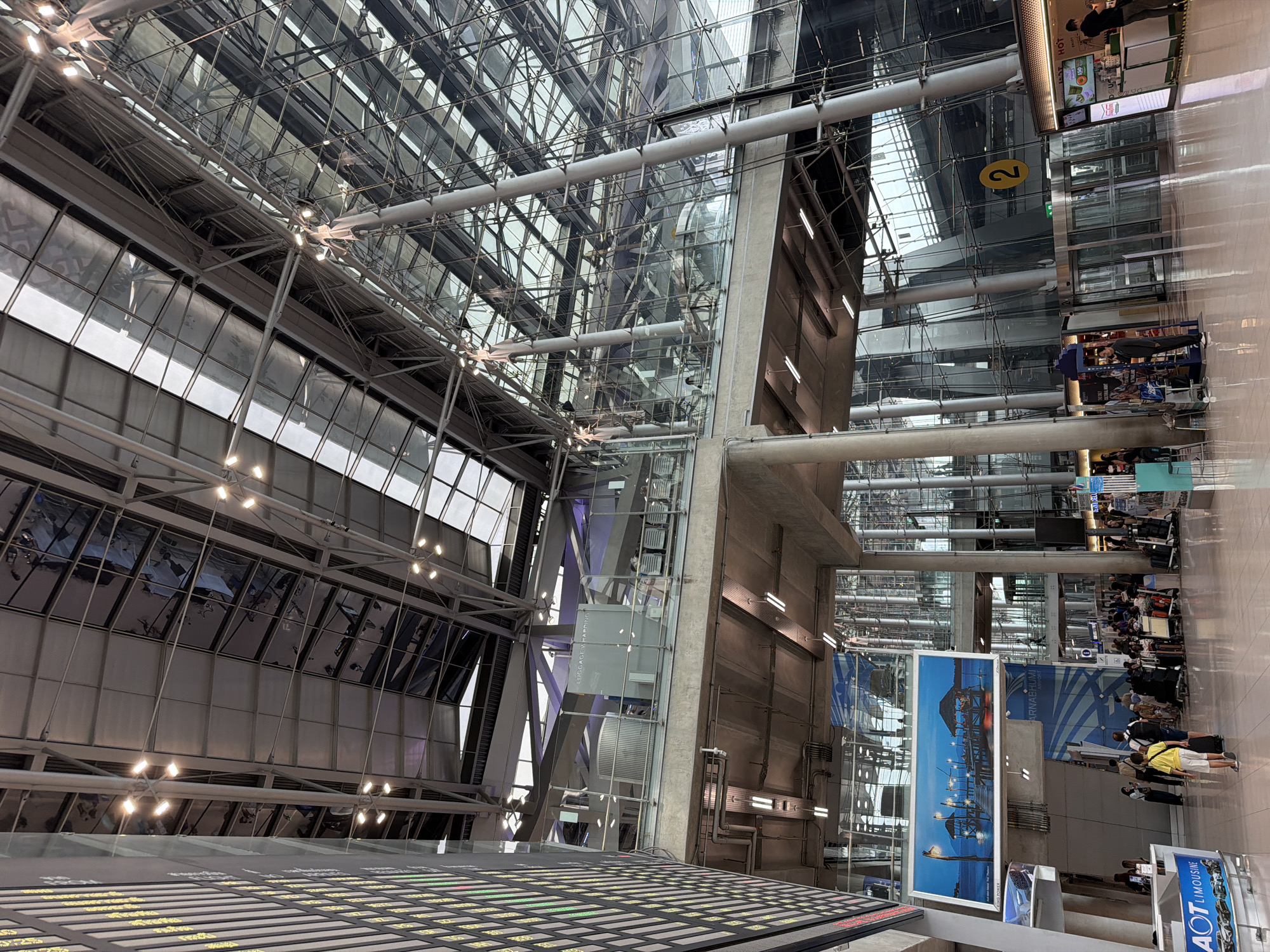 The image shows the interior of a large, modern airport terminal, likely Suvarnabhumi Airport, located in Nong Prue Subdistrict, Thailand. The scene is characterized by its expansive, high-ceilinged architecture with a prominent use of glass and metal structures. Sunlight streams in through a large, multi-paneled glass roof and walls, reflecting off the polished floor.
On the left side of the image, a large electronic display board with rows of yellow and green text is visible, likely showing flight information. Below this, an "AOT Limousine" sign is displayed on a blue background with images of cars.
In the mid-ground, several people are seen walking and standing with luggage. There are seating areas and various service counters or shops. A prominent sign reads "LUGGAGE WRAPPING." A large framed picture depicting a tranquil scene of piers and huts at dusk is hung on a wall.
Further into the terminal, a large crowd of people with trolleys full of luggage suggests an active travel environment, likely near check-in counters or baggage claim. A yellow circle with the number "2" is prominently displayed on a pillar, possibly indicating a gate or section.
The architectural style emphasizes open spaces, transparency with extensive glass facades, and structural beams and columns supporting the roof and upper levels. The overall impression is one of a busy, functional, and architecturally impressive transportation hub.