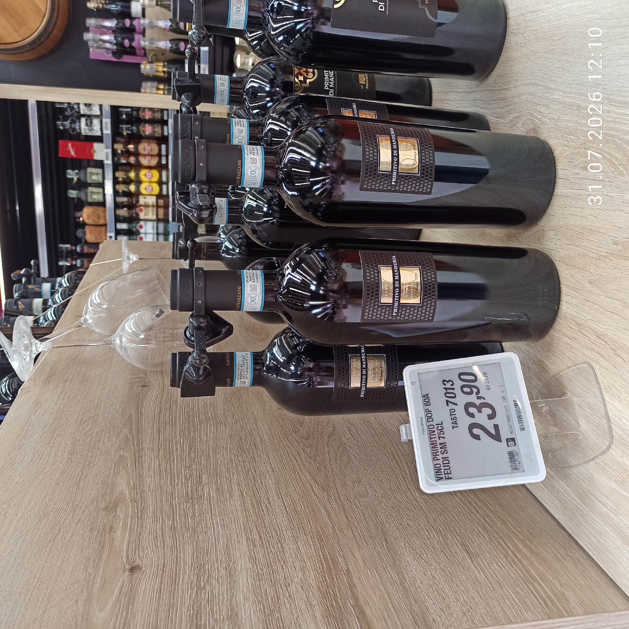 The image shows a close-up view of several bottles of red wine, likely on display in a store. The bottles are dark colored and have gold and black labels. One price tag is visible in the foreground, displaying "VINO PRIMITIVO DOP 60A FEUDI SM 75CL" and a price of "23,90". In the background, shelves are stocked with various other bottles, some of which appear to be champagne or sparkling wine. The overall environment suggests a retail setting, possibly a supermarket or a specialty wine shop. The date stamp at the bottom right corner indicates July 31, 2026, at 12:10 PM. The image does not provide any specific information about the city of Modugno, Italy.