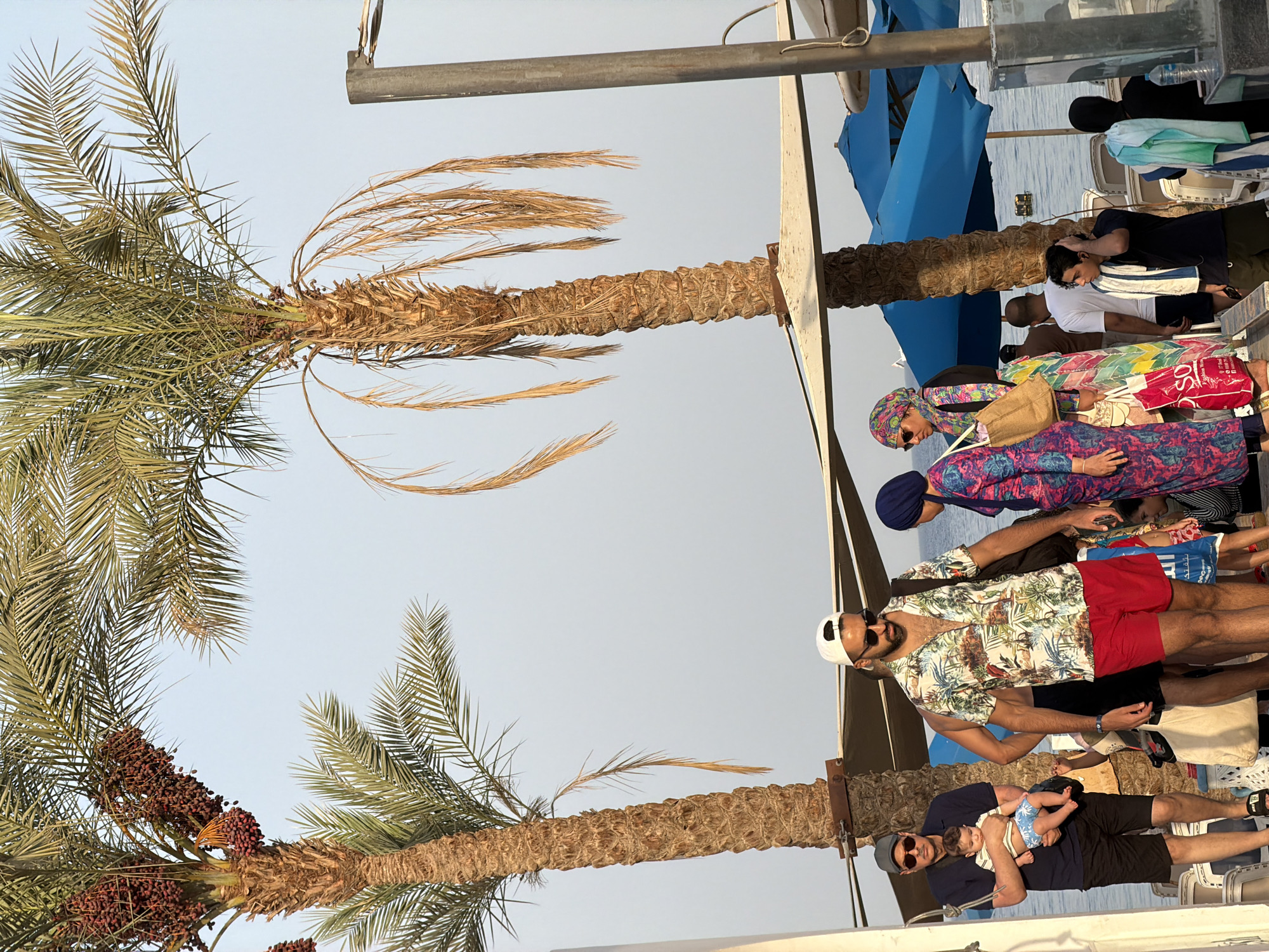 The image depicts a beach scene in Sharm El Sheikh, Egypt, with palm trees and beach umbrellas visible. In the foreground, a group of people are walking. One man in a floral shirt and red shorts is walking towards the viewer, wearing a white baseball cap and sunglasses. To his left, an older man in a dark polo shirt and black shorts carries a baby wearing a striped onesie. Other individuals, some wearing headscarves, are also present, along with beach bags and towels. The background shows the Red Sea under a clear sky.