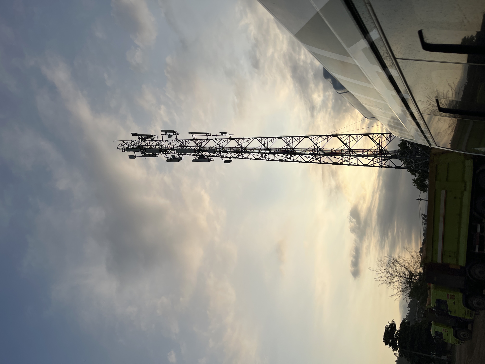 A cellular tower stands tall in the foreground against a cloudy sky at dusk. In the lower portion of the image, two lime-green dump trucks are parked side-by-side. A white vehicle, possibly a van or bus, is positioned to the right, partially obscuring the view of the trucks and the cellular tower. The scene appears to be set in an outdoor industrial or construction area, with hints of trees and hills in the background. The overall lighting suggests the late afternoon or early evening.