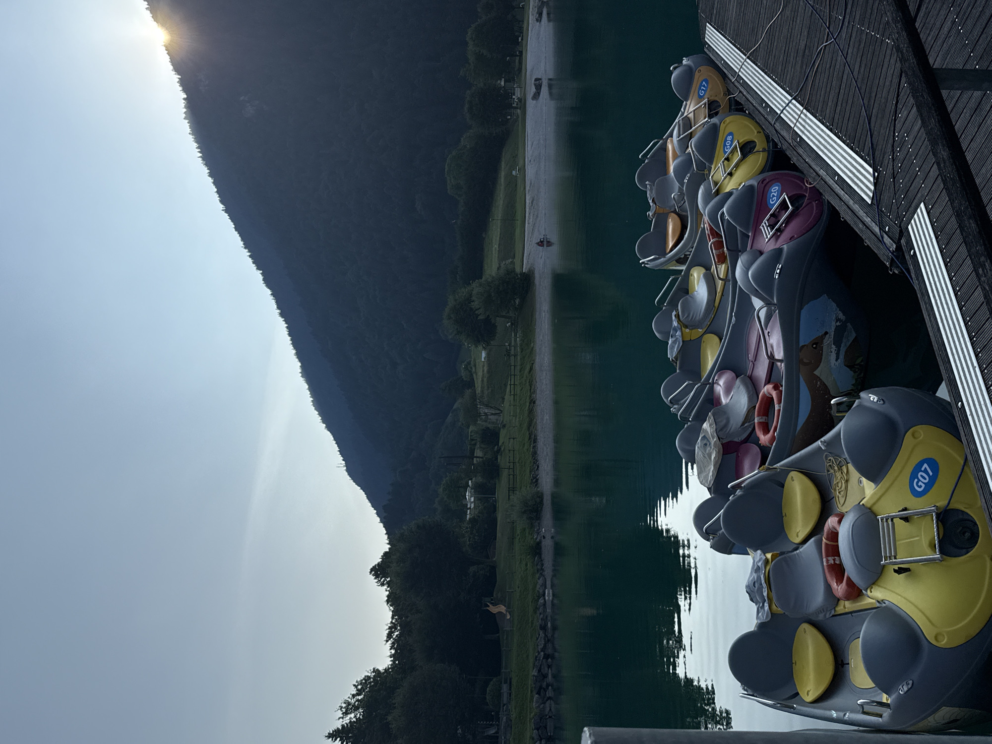 The image depicts a serene scene at Lake Molveno, Italy, captured during a late afternoon or early evening. In the foreground, several pedal boats are docked side-by-side on a wooden jetty. These boats, primarily in shades of gray, yellow, and pink, feature multiple seats and life preservers. Some of the boats are marked with alphanumeric identifiers such as "G07," "G08," and "G17." The water of Lake Molveno is calm and reflects the surrounding environment.
In the middle ground, a beach area is visible, with a few scattered logs and what appears to be a small playground structure. The shoreline is lined with trees, creating a verdant backdrop. Further in the distance, a gentle slope covered in dark green conifer trees rises to form a mountain. The sun is setting behind the mountain, casting a warm glow and creating a sunburst effect where its rays break through the treeline. The sky above is a soft gradient of blue, transitioning from a lighter hue near the horizon to a deeper shade overhead. The overall atmosphere is peaceful and tranquil.