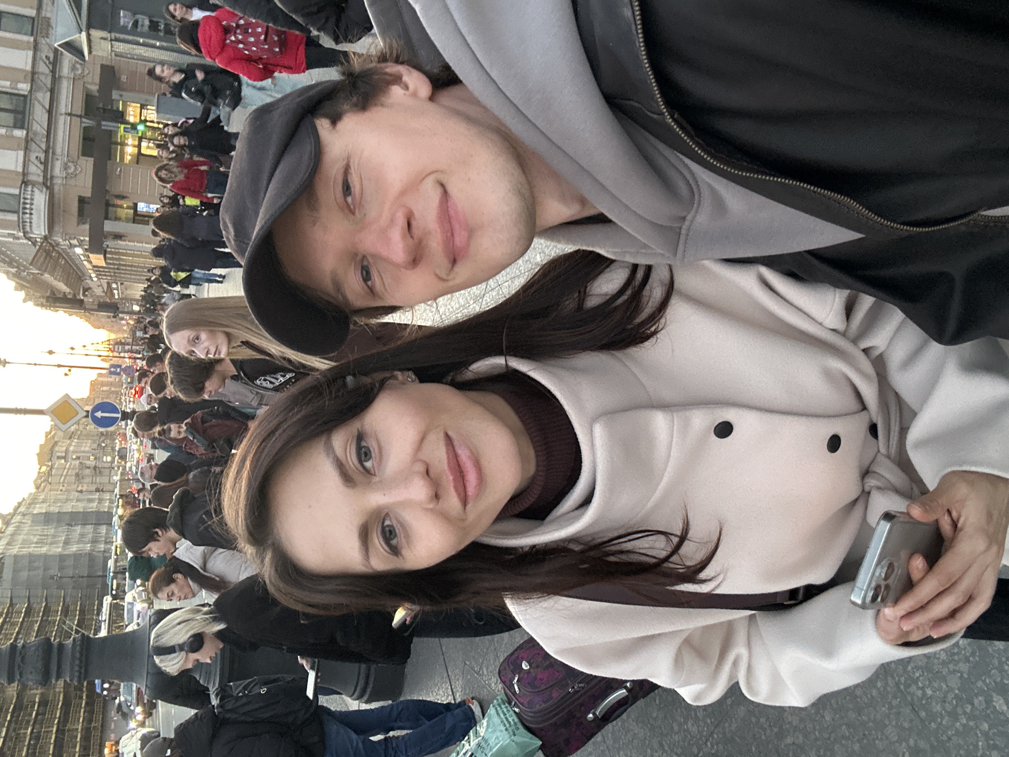 A selfie captures a man and a woman amidst a bustling street scene. The man, sporting a dark baseball cap and a gray hoodie partially obscured by a black jacket, stands slightly behind the woman. The woman, wearing a tan overcoat over a brown turtleneck, holds a smartphone in her hand. They are both smiling at the camera.
Behind them, a crowded street extends into the distance, lined with cars and pedestrians. Street signs are visible, mounted on a light post. The architecture suggests that they are in a city. Luggage is visible on the ground. The image seems to be taken on Nevsky Prospect in Saint Petersburg, Russia.