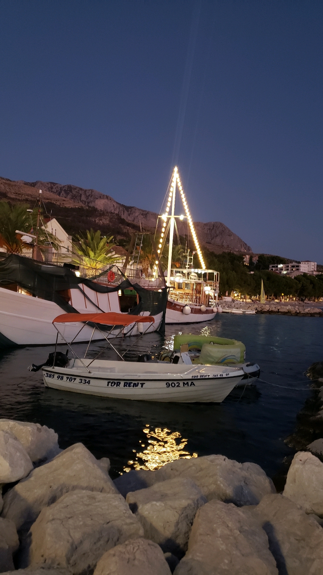 The image depicts a nighttime scene at a harbor in Općina Tučepi, Croatia. The sky is a deep blue, indicating dusk or early night. In the foreground, large rocks are piled up along the water's edge. Several boats are moored in the water. The closest boat is a small white motorboat with an orange canopy, labeled "FOR RENT" and "902 MA". Behind it, a larger white boat is partially visible, with its hull covered by a dark tarp. Further in the background, a tall sailing vessel is illuminated by a string of decorative lights that form a triangular shape up its mast. This vessel has multiple decks and appears to be designed for passengers. To the right of the sailing vessel, more boats are docked along a shoreline that is lined with buildings and palm trees. In the distance, mountains rise up behind the town. The water reflects the lights from the boats and the shore.