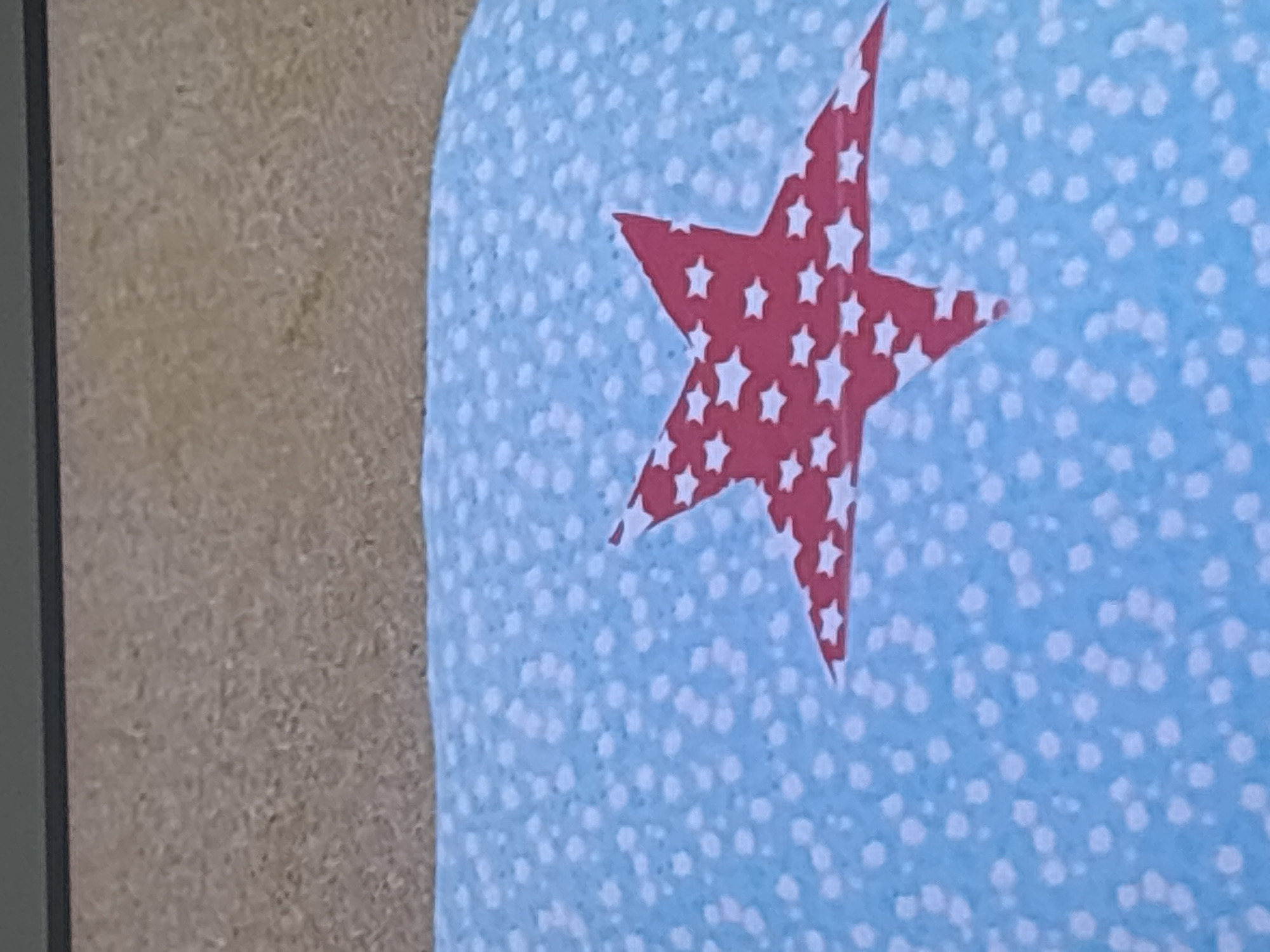 The image displays a close-up view of a light blue fabric with a pattern of white dots and a large red star. The red star is filled with smaller white stars. The fabric appears to be part of a cushion or similar textile item. Above the fabric, a textured surface, possibly wood or a similar material, is visible, with a dark horizontal line separating it from the fabric. The scene is not located in Dubai, United Arab Emirates. There are no people or actions depicted in the image.
