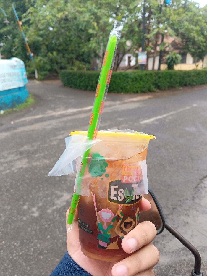 A close-up, eye-level shot shows a person's right hand holding a plastic cup of iced tea outdoors in Cijeungjing, Indonesia. The hand, partially covered by a blue sleeve, grasps the transparent cup, which contains a dark brown liquid and appears to have ice. A vibrant green plastic straw, still in its clear wrapper bearing the words "PULL" and "POCH" in red, is inserted into the drink. A practical, clear plastic carrier is taped to the side of the cup, designed for easy handling.

The cup itself is branded with a colorful label featuring cartoon illustrations of a figure with a pink head and green body, and a brown, bear-like character. Prominently displayed text on the cup includes "TEH POC" in a yellow banner, large white letters spelling "ES T", and "Es T" alongside a leaf icon, with "Est. 1954" also visible. Smaller text, "estehpoci.id" and "estehpoci.co.id", is present near the bottom of the label. Part of a motorcycle's rearview mirror is visible beneath the person's hand, suggesting they may be on or next to a motorcycle.

The background is blurred, showing an asphalt or concrete path lined with green trees and manicured bushes. Distant buildings are discernible through the foliage, and a distinctive blue and white structure is partially visible on the far left. The lighting is soft and diffused, indicating it is daytime, likely under overcast skies or during the early morning/late afternoon, with no harsh shadows present. The image captures a moment of refreshment with a popular local beverage in a casual outdoor setting.
