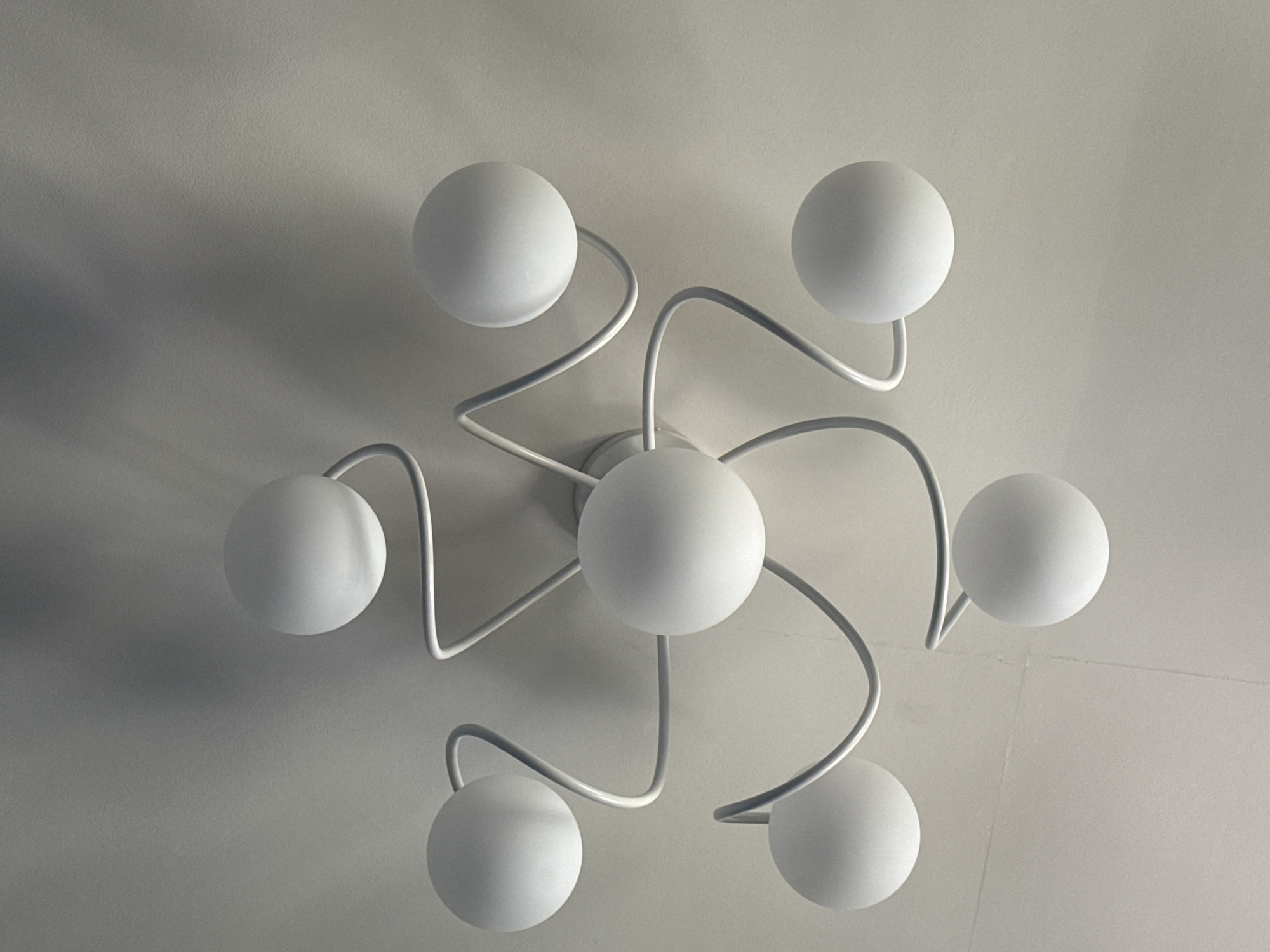 The image shows a white, modern ceiling light fixture with multiple spherical bulbs arranged in a starburst pattern. The fixture is mounted on a textured white ceiling. The lighting is soft and diffused, casting subtle shadows. The style of the fixture suggests it could be a contemporary or minimalist design. There are no people or any other objects visible in the image, and the environment appears to be an indoor space. The image does not provide any information about the city of Rzhev, Russia, or any specific location within it.