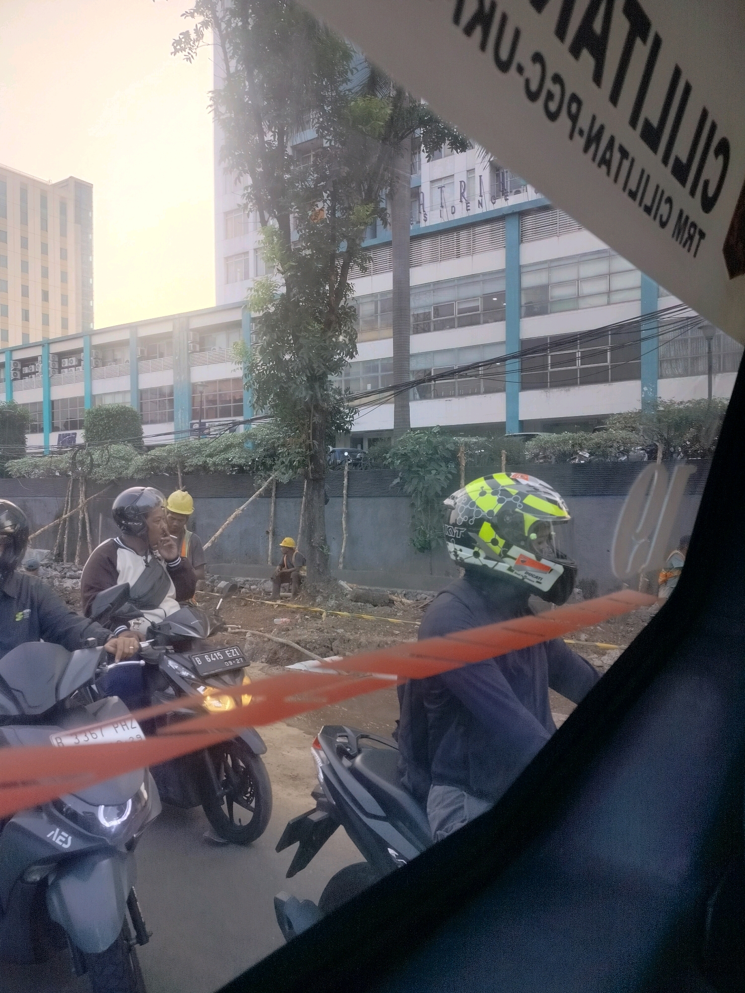 This image depicts an outdoor scene, likely a street in East Jakarta, Indonesia, characterized by construction and urban elements. In the foreground, several motorcycles are parked or in motion. One motorcycle, a dark color with a license plate reading "B 6415 EZ," has its headlights illuminated. Another motorcycle, a lighter shade, has a license plate "B 3367 PHZ."
Several individuals are visible. Two men in the middle ground appear to be construction workers, one wearing a yellow hard hat and a safety vest, the other in a darker shirt. One of them is smoking. Further in the background, another individual in a yellow hard hat is seated. In the right foreground, a person on a motorcycle is wearing a distinctive yellow and black helmet with "KYT" and "DUCATI" branding.
The environment suggests an ongoing construction project, with dirt, debris, and wooden supports visible on the ground. A tall, modern building with many windows forms the backdrop, with some trees interspersed. A banner or sign, partially visible at the top of the image, displays text in a foreign language, likely a warning or informational sign related to the construction or an adjacent facility. The overall lighting suggests it is either early morning or late afternoon, with a bright sky.