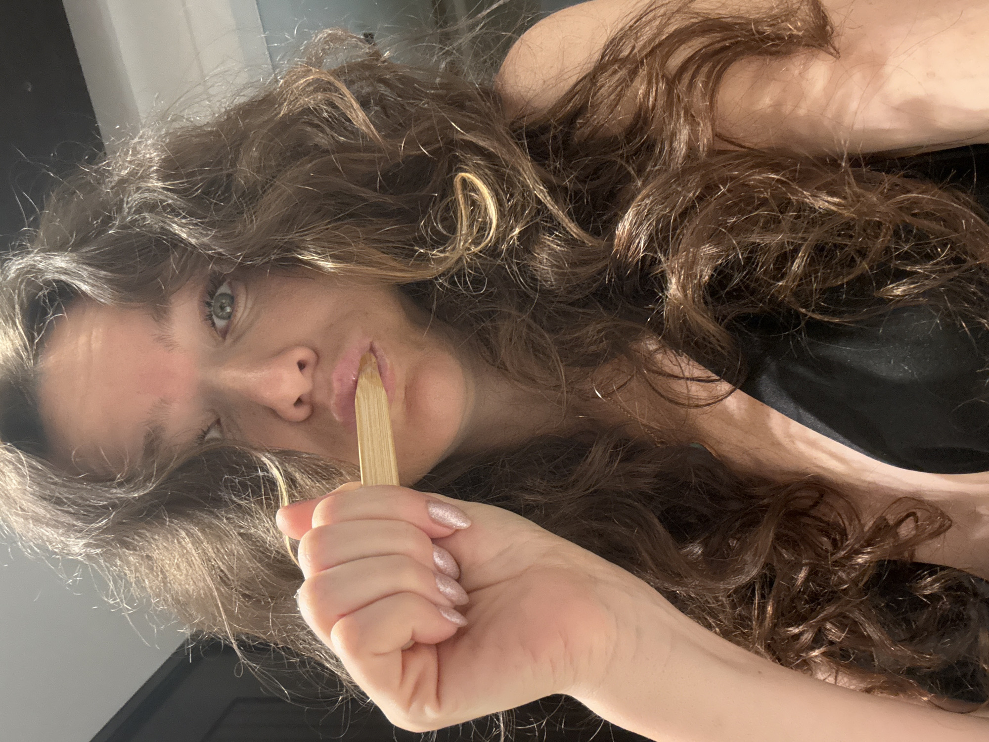 An adult female is depicted in a close-up, selfie-style photograph indoors. She possesses long, wavy brown hair with lighter highlights, framing her face. Her left eye, light in color, is open and directed towards the camera, while her lips are pursed around a light-colored, flat wooden stick held horizontally. Her visible left hand, adorned with sparkling pink nail polish, grasps the stick. She is wearing a black top, visible around her neckline and shoulders. The background is an indistinct indoor setting, featuring a dark surface on the left and a lighter area on the right, suggesting a plain wall or ceiling. There are no identifiable elements within the image that indicate a specific location in the city of Dubai, United Arab Emirates.