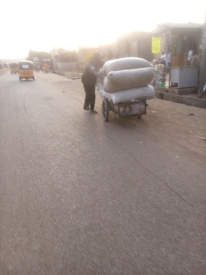 The image captures a vibrant street scene in Rangaza, Nigeria, likely during late afternoon or early morning due to the warm, hazy sunlight filtering through the background.
The central subject is a person, dressed in dark clothing, pushing a two-wheeled handcart laden with three large, light-colored sacks or bales. The cart appears to be moving from left to right along the side of a paved road.
In the mid-ground on the left, several yellow auto-rickshaws, commonly known as Keke Napeps, are visible on the road, suggesting ongoing traffic or a busy thoroughfare. The road surface itself is asphalt, showing some cracks.
On the right side of the frame, alongside a sidewalk, are several informal commercial establishments or market stalls. People are seen near these stalls, some seated. One stall features a small white refrigerator or cooler, and various items are displayed for sale. A yellow sign hanging from an awning over one of the stalls shows blurry text, possibly "20N" or a similar numerical indicator, likely a price or denomination.
The overall setting is characteristic of a bustling urban or semi-urban area in Nigeria, with a mix of street vendors, local transportation, and daily activity under a bright, diffused light. No strong emotions are discernible from the individuals, but the scene conveys a sense of everyday commerce and movement.
