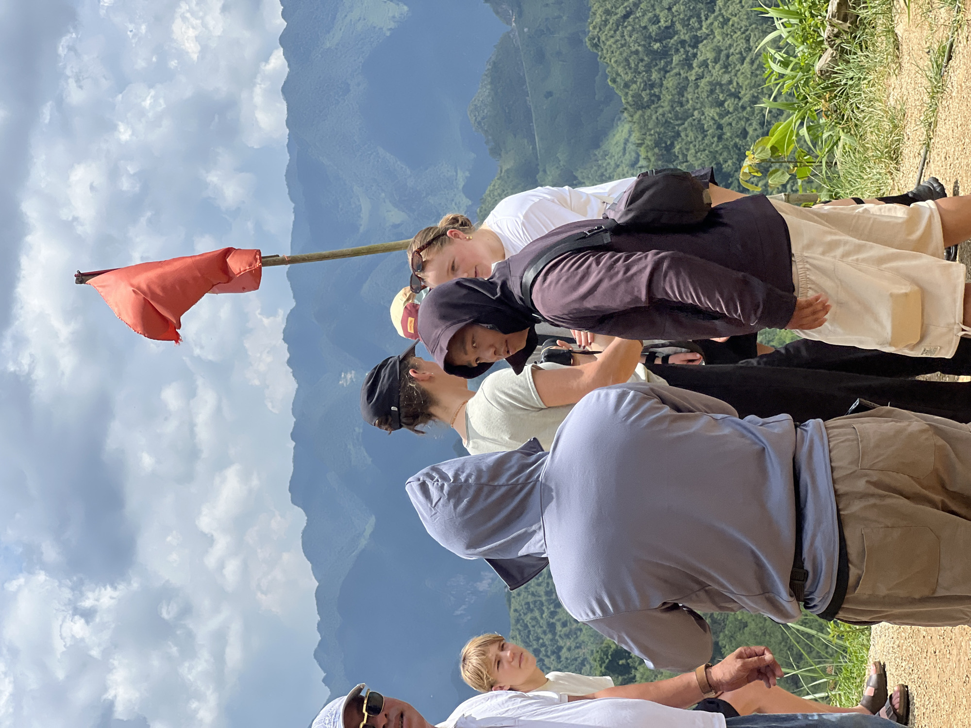 A group of people are gathered outdoors on a dirt path with a lush, mountainous landscape in the background under a cloudy sky. A red flag on a wooden pole is prominent in the upper right of the frame. The individuals, facing away from the camera and towards the landscape, are dressed in casual, outdoor attire including hooded jackets and shorts. One person in the foreground wears a grey hooded jacket and tan shorts. Another person to their left wears a dark hooded jacket and light-colored shorts. A young boy with blonde hair is visible to the left, looking towards the group. Further back, a woman in a white t-shirt and sunglasses is speaking with others. The overall impression is of tourists observing a scenic view. The location is the Heaven's Gate viewpoint in Quản Bạ Commune, Vietnam.