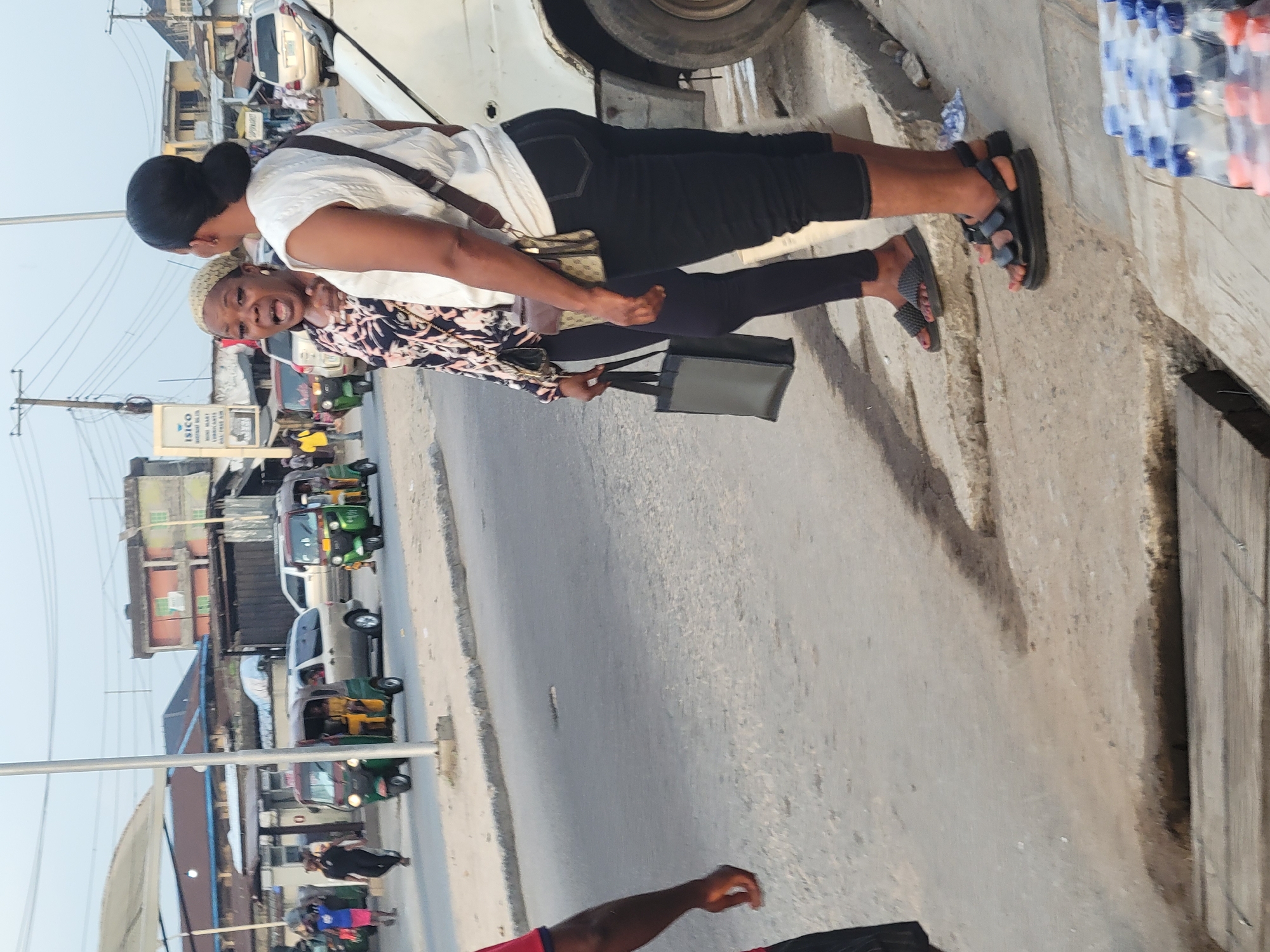 The image shows two women standing on a sidewalk next to a street in Aba, Nigeria. The woman in front wears a patterned shirt, black leggings, and sandals, carrying a black bag. The woman behind her wears a white t-shirt, black pants, and sandals, with a small shoulder bag. The street has auto rickshaws (tuk-tuks) and other vehicles. A sign for "ISICO" is visible in the background. Bottles of water are stacked nearby. The environment appears to be a busy urban commercial area.