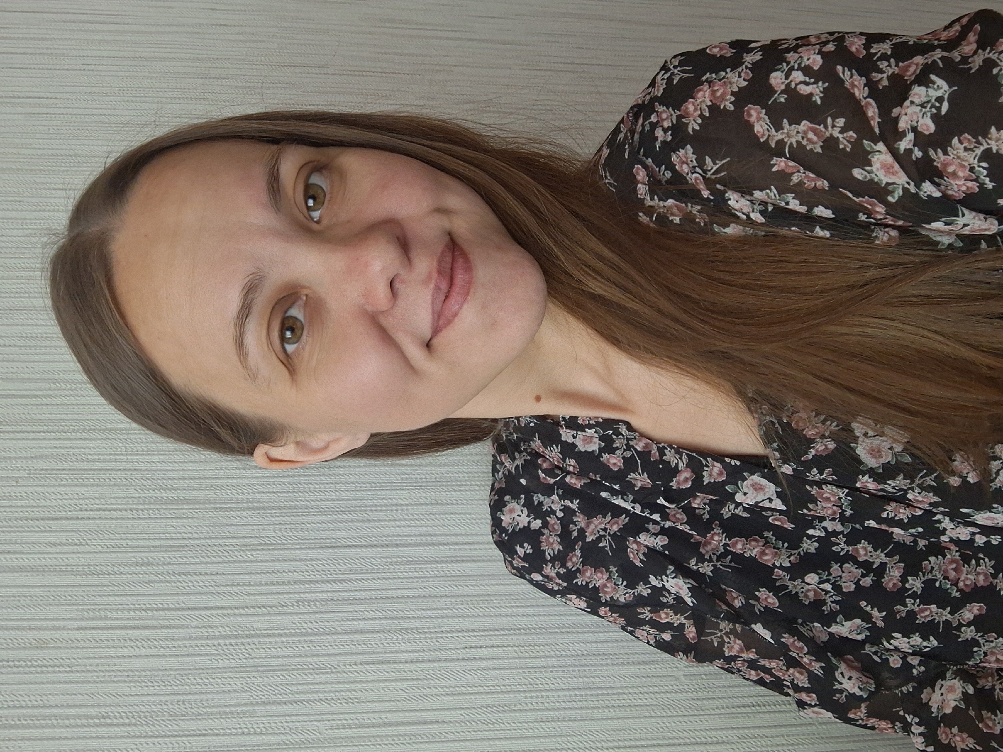 A light-skinned woman with brown hair and eyes is pictured from the chest up. She is wearing a black floral dress with pink and white flowers. Her hair is straight and falls past her shoulders. She is looking directly at the camera with a slight smile. The background is a neutral-colored wall with a subtle horizontal pattern. The location is likely indoors but cannot be confirmed as being in Chaikovsky, Russia, based on the visual information alone. There are no discernible actions or interactions visible.