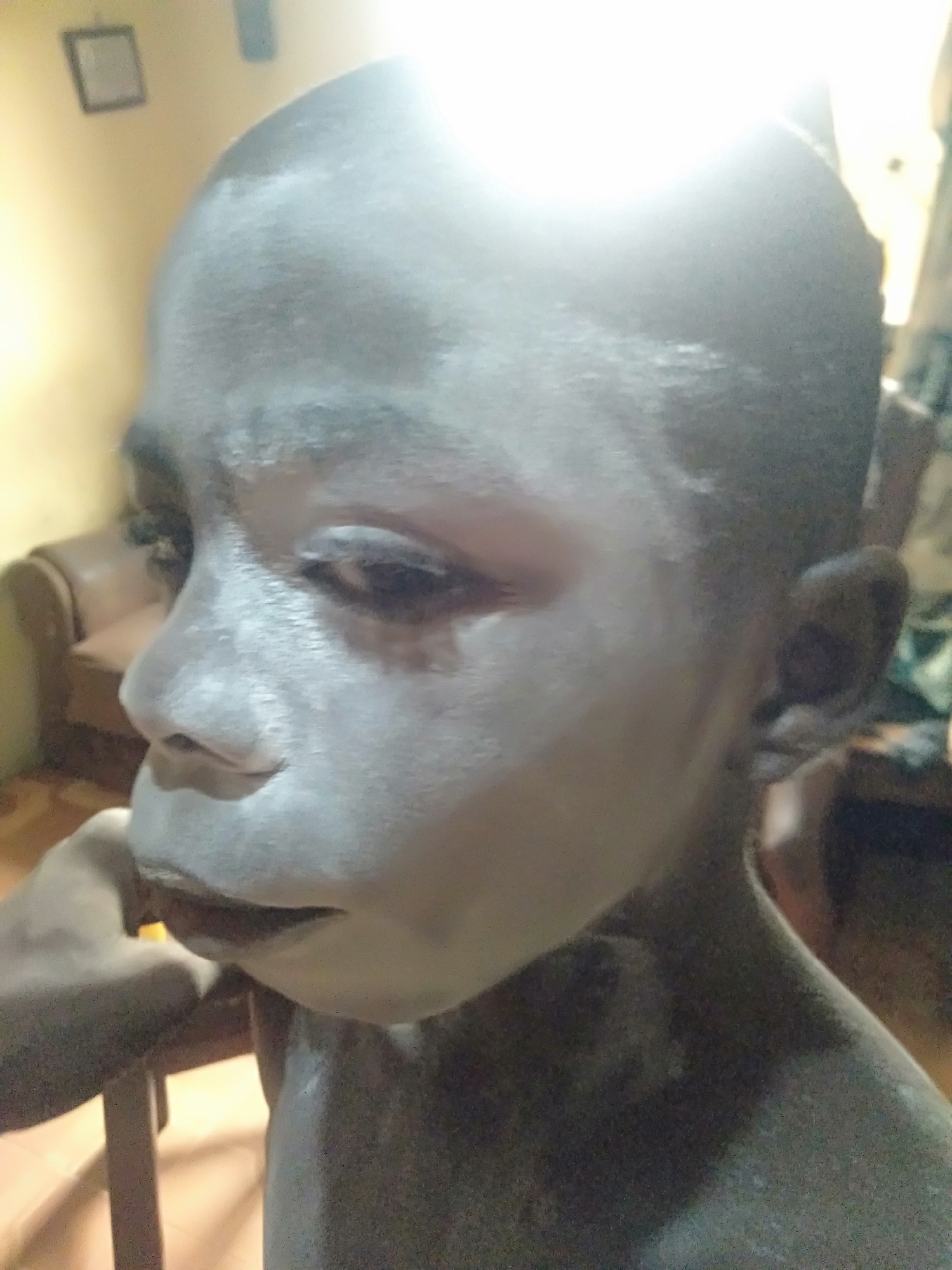 A close-up, slightly out-of-focus photograph depicts a child's face and upper body from a low-angle perspective, possibly in Shangisha, Nigeria. The child's skin is covered in a white substance, likely paint or powder, which obscures the natural skin tone. The face is turned to the side, with the eyes appearing downcast or closed. The child's right ear is visible. A hand, likely belonging to someone else, is near the child's face. The background is a blurred indoor setting, with a glimpse of furniture and a framed picture on the wall. The lighting is bright and somewhat harsh, creating a glare on the child's forehead.