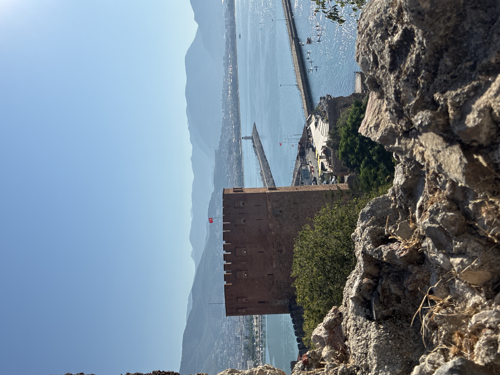 The image shows a view of the Red Tower (Kızıl Kule) and the harbor in Alanya, Turkey. The Red Tower, a prominent historical landmark, is a tall, octagonal brick structure with crenellated battlements, flying a Turkish flag. It is situated on a rocky outcrop overlooking the Mediterranean Sea.
In the background, the city of Alanya stretches along the coastline, with buildings nestled against rolling, hazy mountains. The sea is a vibrant blue, dotted with several boats and a long pier extending into the water. A lighthouse stands at the end of one of the piers. Several people are visible on the water, some on paddleboards.
The foreground is dominated by weathered, rocky terrain, suggesting the viewpoint is from a similar historical fortification or natural elevation. The sky is clear and a bright, uniform blue, with a hint of sun glare in the upper right corner. The overall scene depicts a sunny day in a coastal city known for its historical sites and tourist attractions.