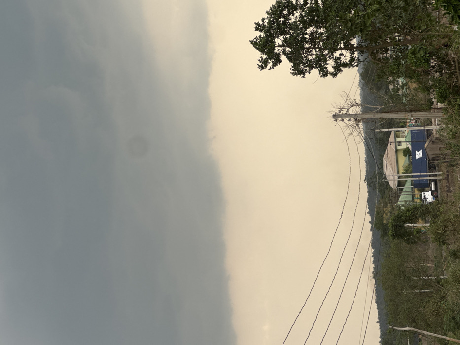 This image captures a semi-urban or rural landscape in Ipetumodu, Nigeria, under an overcast sky. The upper approximately 40% of the frame is dominated by heavy, dark grey clouds, while the lower portion of the sky appears a lighter yellowish-brown, indicating atmospheric haze or specific lighting conditions.
In the foreground, four dark, parallel utility wires stretch horizontally across the lower-mid section of the image from left to right. To the right, a weathered wooden utility pole supports these wires, with additional tangled wires and bare branches extending from its top. Immediately to the right of the pole, a large, leafy green tree partially obscures the view. The ground directly beneath the utility lines and in the mid-ground is rough, unpaved terrain with sparse, low-lying green vegetation and brown patches of earth.
In the mid-ground, a white truck with a blue shipping container is visible, positioned facing towards the left. The container features a white "K" or "K Line" logo on its side. Behind the truck, a single-story building with light yellow walls and a light yellow-green hip roof is prominent. Further back and slightly to the left, another building with a dark green roof is partially visible amidst trees.
The background consists of a range of low hills or elevated terrain, densely covered in dark green foliage, appearing somewhat obscured by atmospheric haze. No individuals are discernible within the frame. The overall environment suggests a mix of natural vegetation and human habitation, with visible infrastructure for utilities and transport.