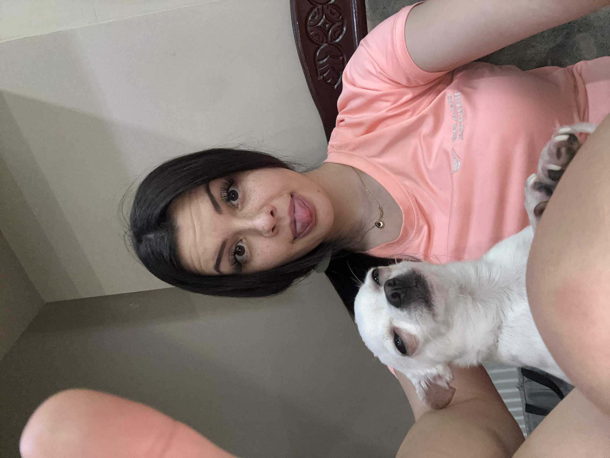 A young woman with dark hair and light skin, adorned with freckles and false eyelashes, is captured in a close-up selfie. She is wearing a pink t-shirt with a metallic logo and a delicate necklace. Her tongue is playfully sticking out, and she is holding a small white chihuahua on her lap. The dog, with its eyes squinted and a somewhat unimpressed expression, is positioned in the foreground, partially obscuring the woman's chest. The background consists of a plain wall and a glimpse of a dark wooden headboard. The scene appears to be indoors, possibly in a bedroom. The image does not contain any information about the city of Sliven, Bulgaria.