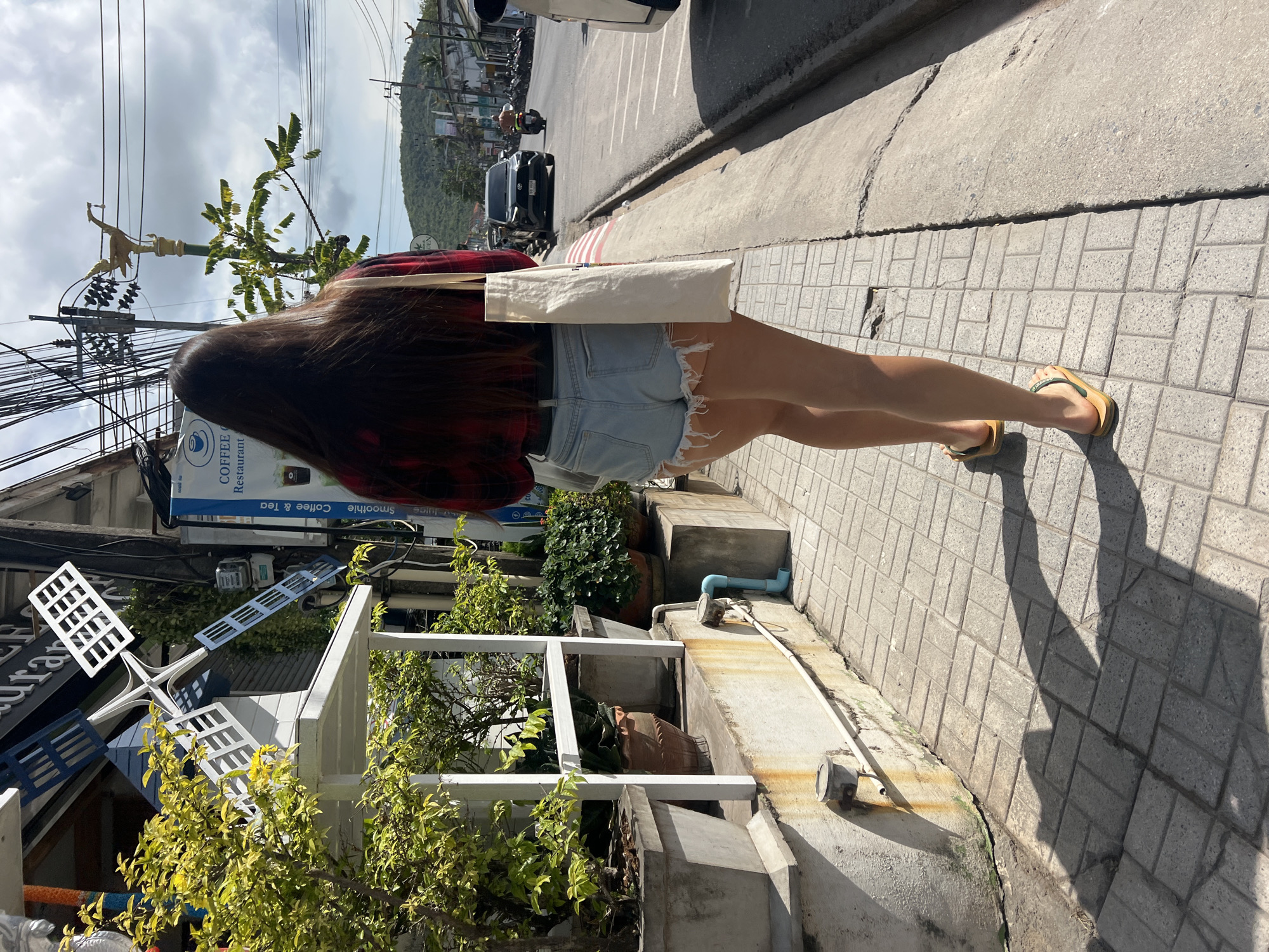 A person walks down a sidewalk in Baan Bang Rak, Thailand. The person has long brown hair, wears a red and black plaid shirt, denim shorts, and yellow sandals. They carry a white tote bag. The sidewalk is made of gray brick. Next to the sidewalk, there is a white, metal railing enclosing plants in pots. On the other side of the sidewalk, there is a street with cars and motorbikes. Buildings and hills are visible in the background. Power lines cross above the scene. A sign with "COFFEE & TEA" is visible on a building.