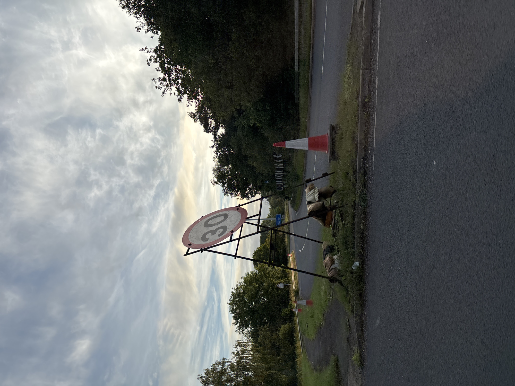 A 30 miles per hour speed limit sign is visible on a metal frame by the side of a road in South Staffordshire, United Kingdom. The sign is circular with a red border and a white background, displaying the number "30" in black. Two traffic cones are placed in front of the sign, one red and white, the other orange. Behind the cones, a collection of objects are piled up, including what appears to be discarded clothing or fabric. The road curves to the left, bordered by grassy verges and trees. The sky is cloudy with hints of sunset.