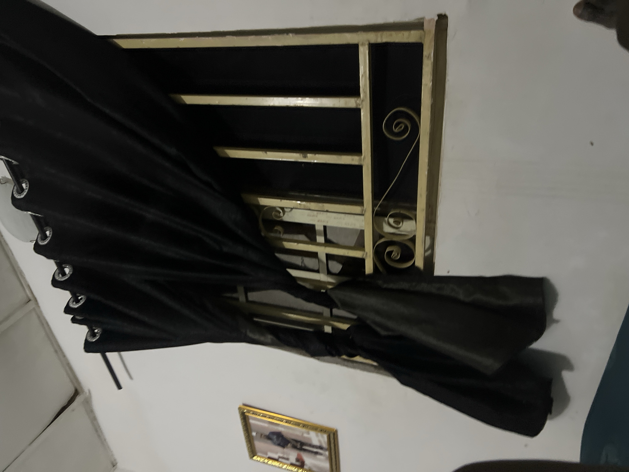 A framed portrait hangs on a white wall, to the left of a barred window covered by a black curtain. The window is ornate, with scrollwork designs on the bars. The room appears to be dimly lit, with the black curtain casting a shadow on the wall. The location is stated to be Howayi, Nigeria.