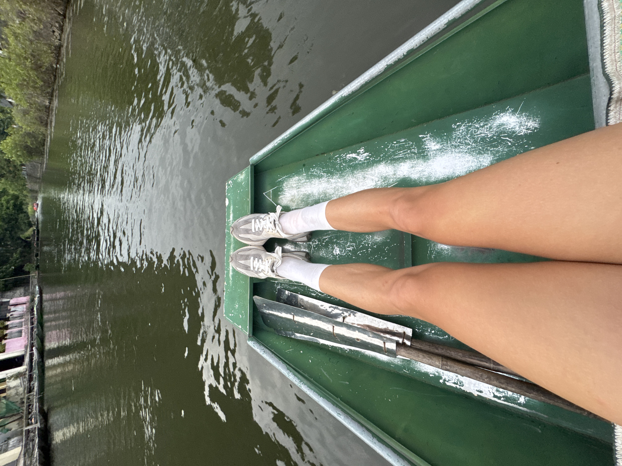 A first-person perspective shot shows a person's legs and feet in a green boat on a body of water. The person is wearing white socks and grey athletic shoes. Two oars with wooden handles and metallic blades are lying inside the boat next to the person's legs. The water is dark green and appears calm, with reflections of the surrounding environment visible on its surface. In the background, a shoreline is lined with buildings and trees. The buildings appear to be of varied colors and architectural styles, with some showing signs of age. The overall scene suggests a leisurely activity, possibly sightseeing or rowing, in a somewhat urbanized natural setting. The location is identified as Nam Hoa Lư Ward, Vietnam.