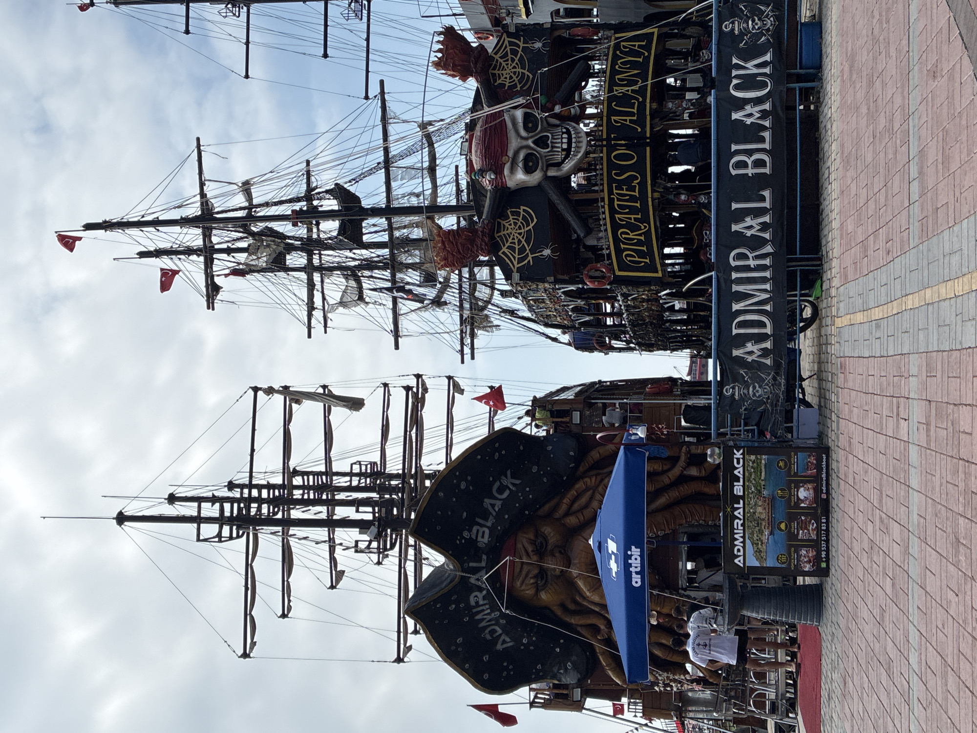 The image shows two pirate-themed boats docked side-by-side in Alanya, Turkey. The boat on the left, named "ADMIRAL BLACK," features a large, ominous kraken-like figure on its stern, wearing a pirate hat. The boat on the right is decorated with a large skull and crossbones, and the text "PIRATES OF ALANYA" is visible on its side. Both boats have multiple masts and rigging, typical of sailing ships. In the foreground, a paved walkway with red and grey bricks is visible. Several people are standing on the dock near the "ADMIRAL BLACK" boat, and a blue umbrella with the "artibir" logo is set up. A banner with "ADMIRAL BLACK" and a smaller image of the boat is displayed on the dock. The sky is overcast with clouds. The overall scene depicts a tourist attraction, likely a pirate boat tour operation in the port of Alanya.