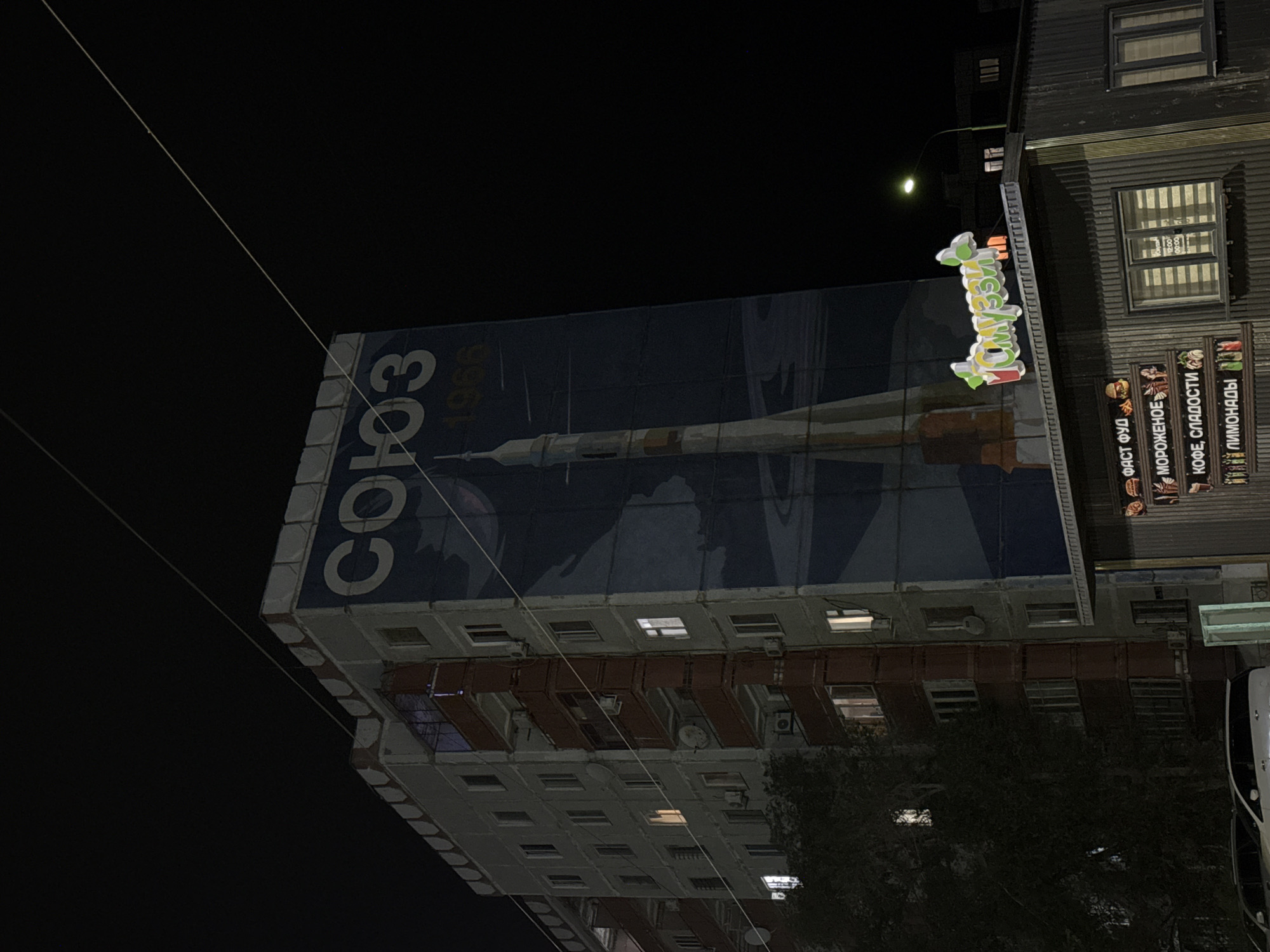 The image depicts a building facade at night, illuminated by the glow of a neon sign and lights from within the building's windows. The building itself appears to be a multi-story structure with a dark, corrugated metal exterior on the upper portion. Below this, there's a section with numerous windows, some of which are lit, suggesting occupancy or activity inside. Reddish-brown balconies or architectural elements are visible between some of the window levels.
A large banner or mural covers a significant portion of the building, featuring a depiction of a rocket and the Russian word "СОЮЗ" (Soyuz) prominently displayed. The year "1966" is also visible on the banner. The style of the banner suggests a historical or commemorative theme.
Attached to the side of the building is a brightly lit, colorful neon sign with Russian text. The sign is stylized and appears to be advertising something. Below the neon sign, on the lower part of the building, there are several smaller signs with text in Russian, indicating businesses or offerings such as "ФАСТ ФУД" (Fast Food), "МОРОЖЕНОЕ" (Ice Cream), "КОФЕ, СЛАДОСТИ" (Coffee, Sweets), and "ПИМОНАДЫ" (Lemonades).
The overall scene is set at night, as indicated by the dark sky and artificial lighting. The location is identified as Baikonur, Kazakhstan. The image is oriented sideways, requiring rotation for a standard view. No people are clearly visible. The mood is somewhat evocative of urban nightlife or a commercial district.