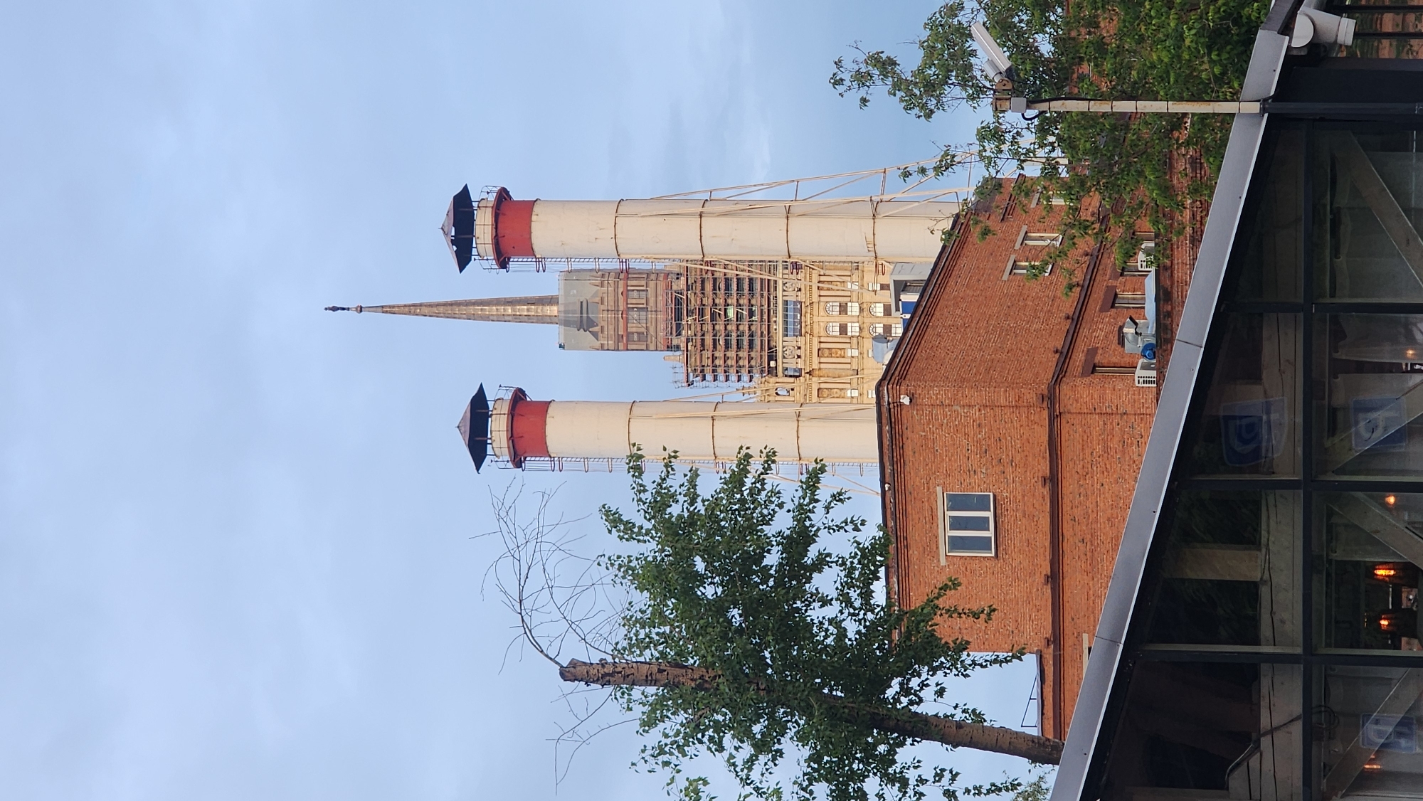 The image features a view of the Kraftwerk Berlin building, located in Moscow, Russia. Two prominent chimneys with red tops and white bodies are visible, extending upwards into a clear blue sky. A church spire is visible between them. Part of a red brick building is to the right. A partially-visible tree with green leaves and bare branches is in the foreground. The corner of another building is also in the foreground. The overall impression is of an industrial and historical landscape within the urban setting.