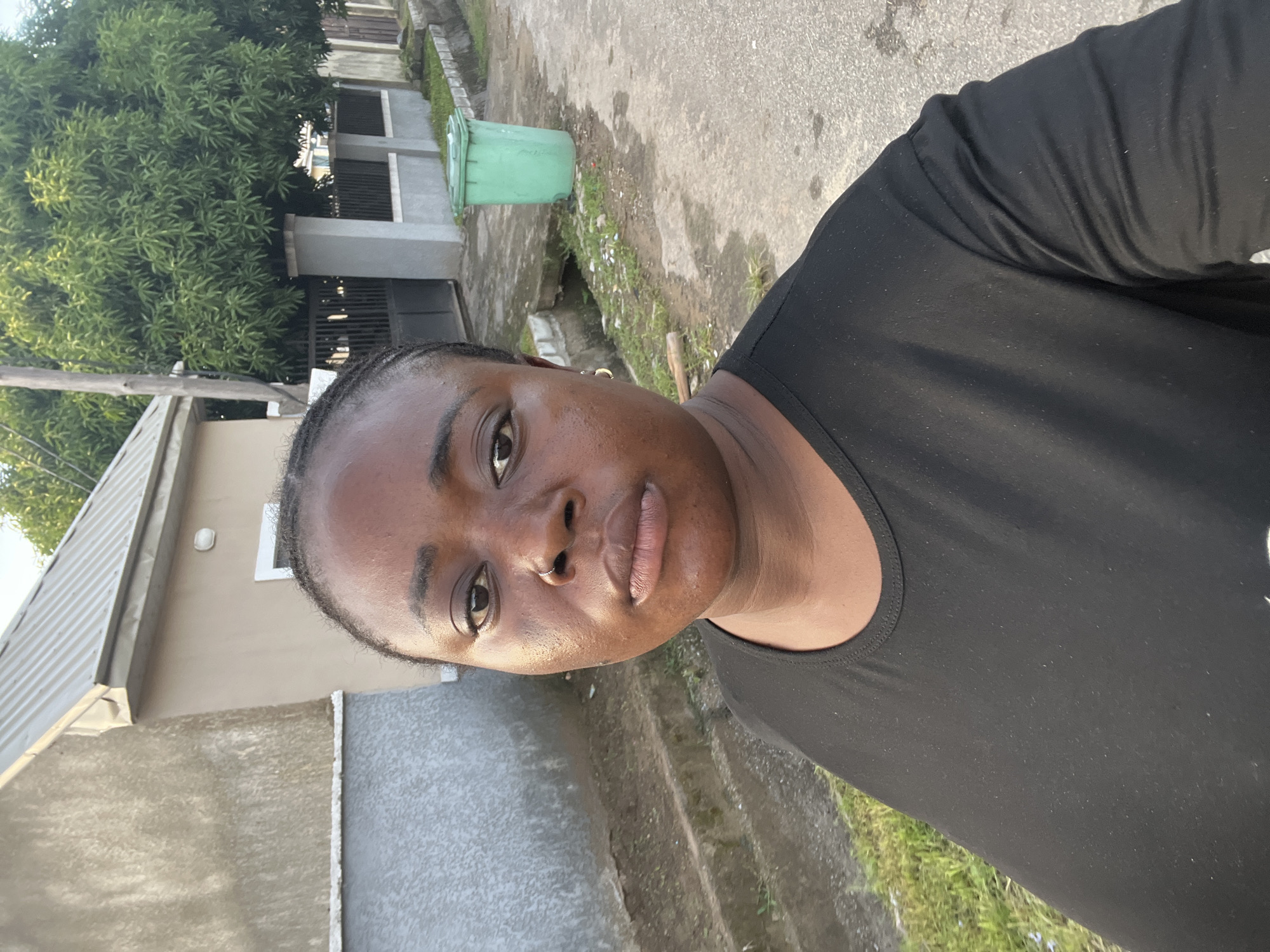 A selfie-style portrait features an adult woman outdoors, facing the camera with a neutral expression. She has dark skin, braided hair pulled back, and is wearing a plain black t-shirt or top. A subtle nose ring is visible on her left nostril.
The background depicts a residential or semi-urban outdoor setting, consistent with the provided location context of Abuja, Nigeria. To the left, a portion of a light-colored building with a corrugated metal roof is visible, along with what appears to be a wooden utility pole. In the mid-ground, a bright green trash bin or bucket stands next to a grey wall or fence structure. A large tree with dense green foliage occupies a significant part of the background behind the fence. The ground is a mix of paved concrete or asphalt and patches of grass and dirt.
The lighting suggests either late afternoon or early morning, with soft, bright sunlight illuminating the scene. The weather appears clear and warm. The overall environment indicates a typical street or compound in a tropical residential area. No visible text is present in the image.