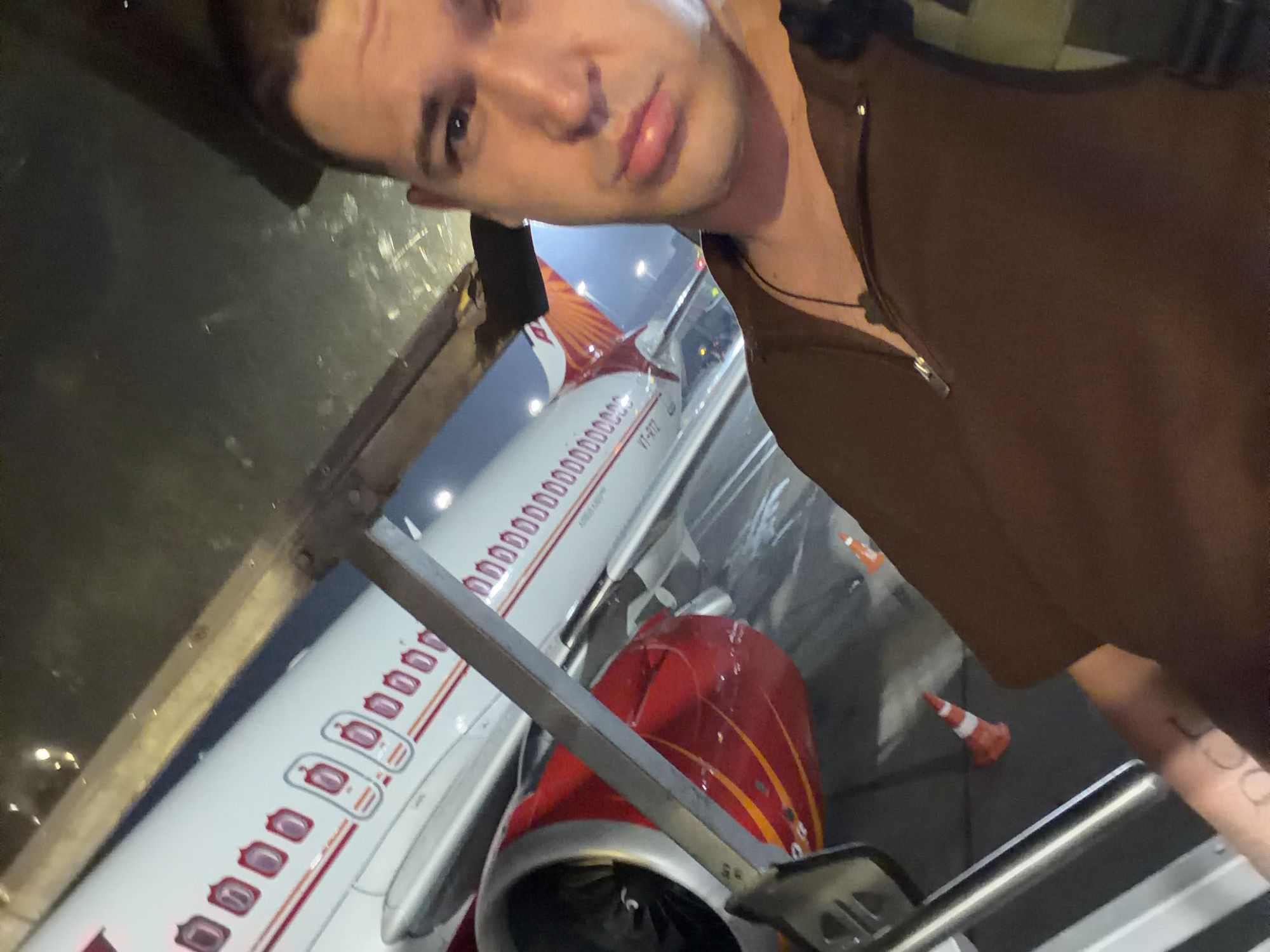 A man is taking a selfie in front of an airplane at an airport. The man is wearing a brown shirt and has a backpack on. The airplane is white with red and orange accents. The image appears to be taken at night or in low light conditions. The location is the Indira Gandhi International Airport in New Delhi, India.