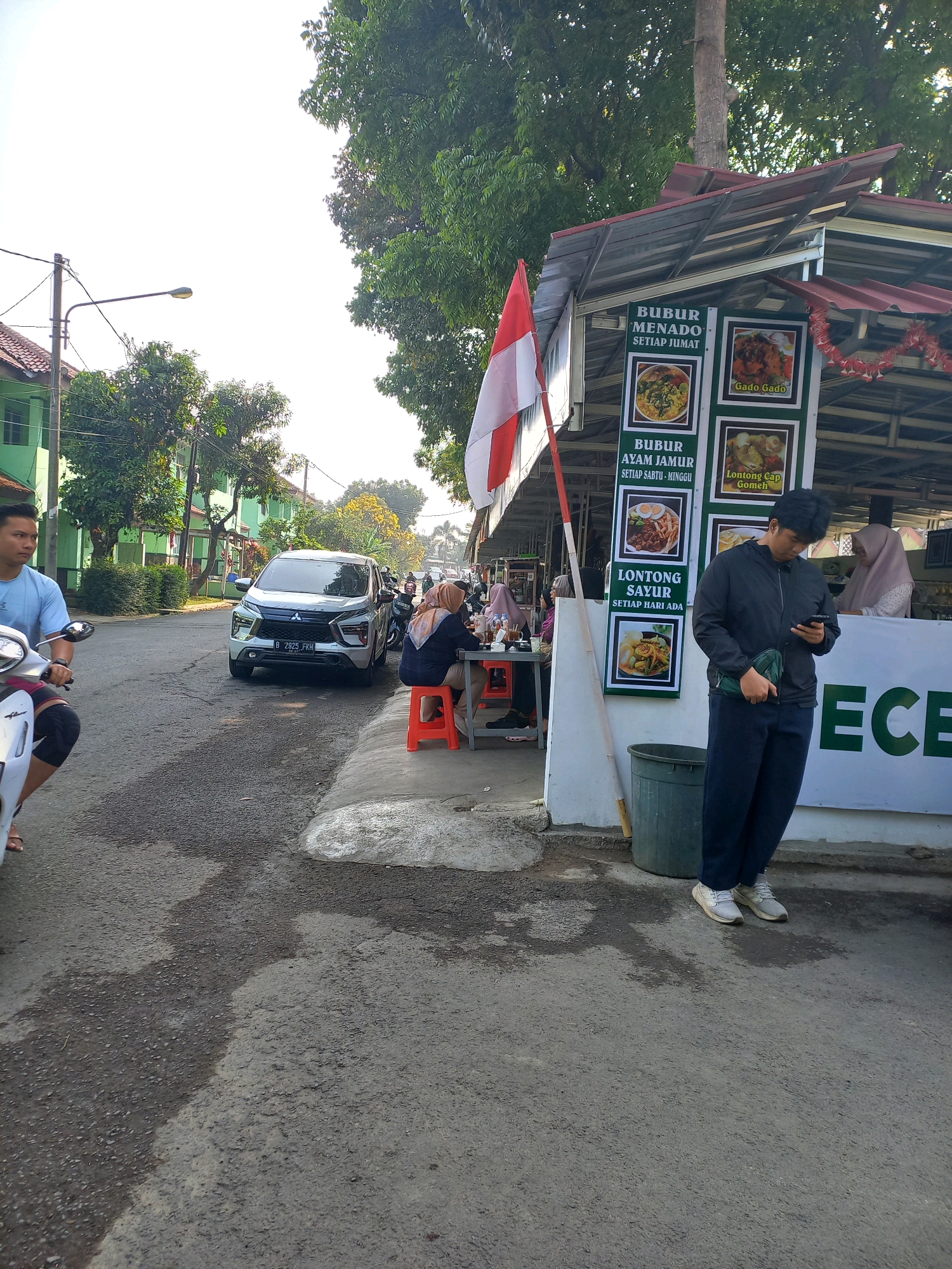 An exterior, medium shot captures a roadside food stall in Cimahi, Indonesia, on a bright, sunny day. The stall features a red, corrugated metal roof and displays various Indonesian dishes on framed posters. In the foreground, a young man in a black jacket and dark pants stands looking at his phone, with a green pouch slung across his body. To his left, a grey SUV with license plate B 2925 FKH is parked on the asphalt road. Further down the road, a motorcycle is visible, with a person wearing a blue shirt on it. Several people are seated at tables in front of the stall, including a woman in a hijab. The environment includes a sidewalk, trees, and residential buildings with green paint.