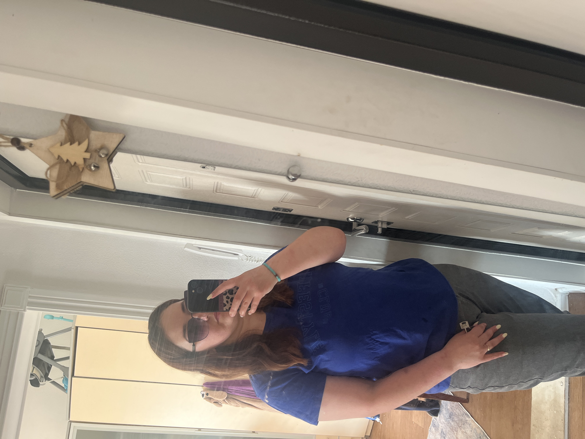 A woman is taking a selfie in a mirror. She is wearing a blue t-shirt, grey pants, and sunglasses. She has long brown hair and is wearing a bracelet on her left wrist. She is also wearing a ring on her left ring finger. In the background, there are doors and a wooden star hanging on the door. There is also a rug on the floor. The scene appears to be indoors.