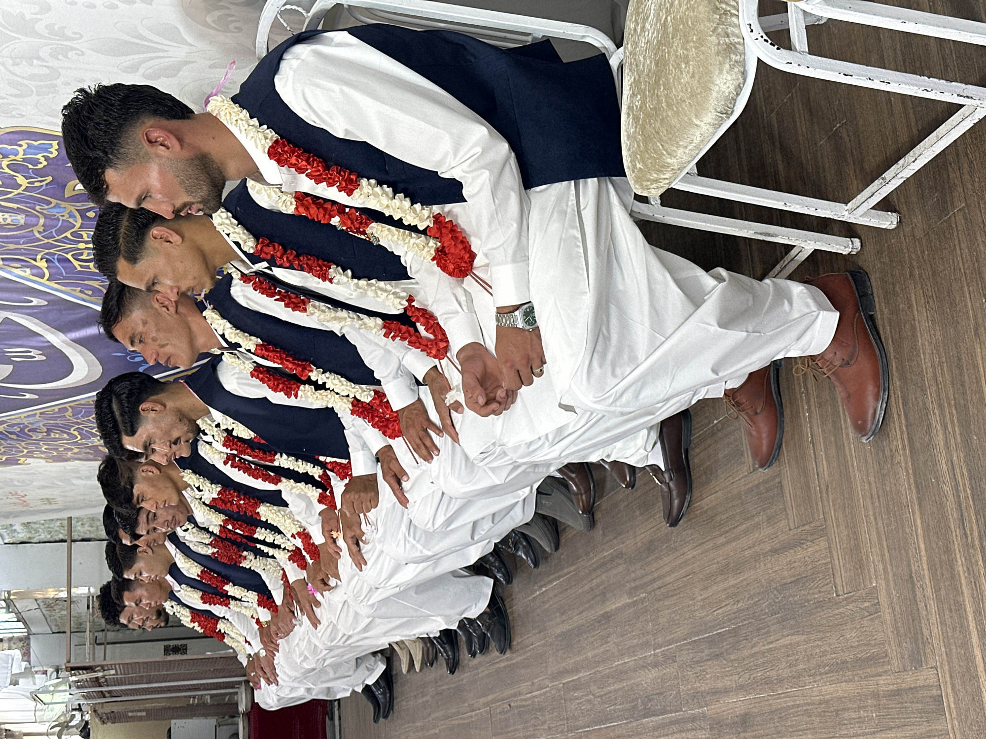 In Quetta City Tehsil, Pakistan, a row of men are seated close together on chairs, likely indoors based on the wooden floor and wall decorations. Each man wears a white shalwar kameez, a navy blue vest, and a garland of red and white flowers. They appear to be part of the same group or event, as their outfits are identical. The men are lined up closely, suggesting a formal gathering or ceremony. The setting appears to be a celebration or special occasion, given the attire of the men and the presence of the garlands.