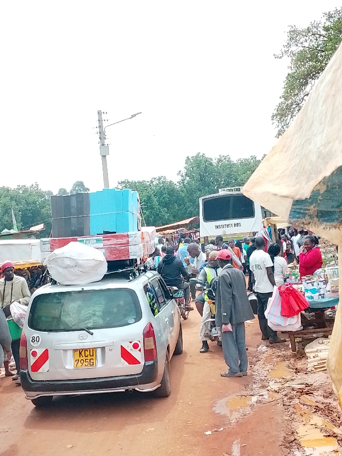 The image shows a busy street scene in Lwandeti, Kenya (latitude: 0.563, longitude: 34.806). A silver Toyota Previa, loaded with goods on its roof rack, is parked on a dirt road with several other vehicles. The car's license plate reads "KCU 7956." There are numerous people walking, shopping, and standing around the car. A white bus with the text "INSTITUTE BUS" visible on its side is parked in the background. Some people are carrying bags and boxes. A few are interacting with vendors at a makeshift market stall with a red and white canopy. The scene is chaotic and colorful, with a mix of people, vehicles, and goods. The road is dusty and covered in muddy puddles. There are trees in the background and a light pole with wires extending from it. The sky is overcast.