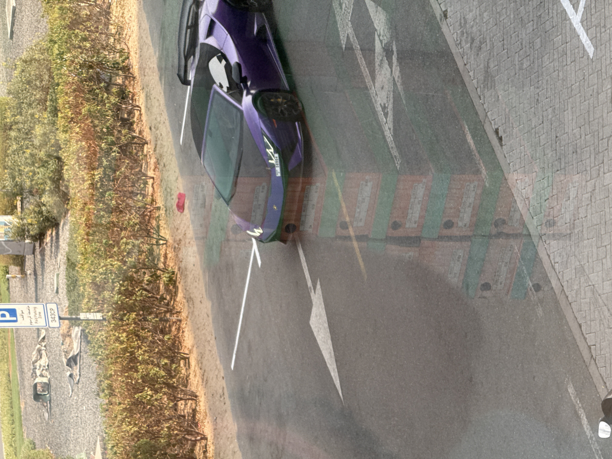 In Dubai, United Arab Emirates, a purple convertible Lamborghini is parked on a paved surface adjacent to a landscaped area with bushes. The vehicle, bearing a license plate with the numerals "38964", is positioned on a road marked with white directional arrows and parking lines. Reflecting on the vehicle's surface are images of stacked boxes, possibly from a nearby building or shipment. In the background, a "Paid Parking Zone" sign is visible next to a gravel area. The overall scene suggests an outdoor urban environment.