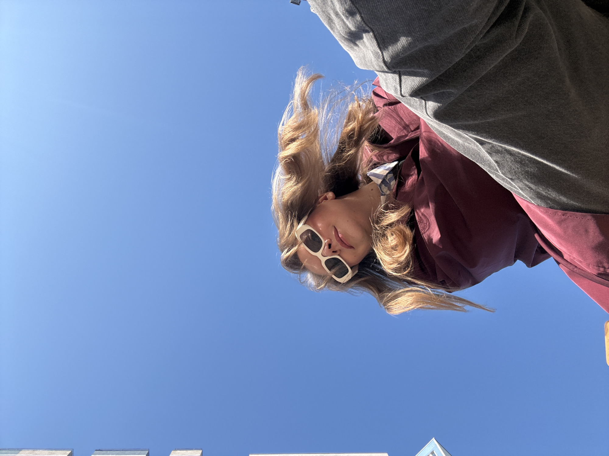 A woman with blonde hair and white-rimmed sunglasses looks down at the camera against a clear blue sky. She wears a maroon jacket partially obscured by a grey top. In the lower portion of the frame, portions of buildings are visible. It is not possible to determine from this image if the location is in the city of Izhevsk, Russia.