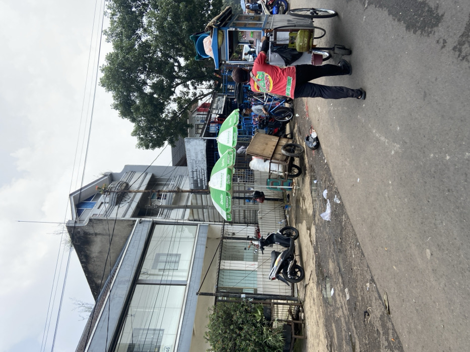 The scene is a street in Bandung, Indonesia, located at coordinates -6.931, 107.602. A man is walking along the street, pushing a food cart. He is wearing a red and yellow shirt with a green design on the back. The food cart is filled with food, and there is a green and white umbrella on the top. Behind him are a few people walking and riding bicycles. In the foreground, there is a black scooter parked on the side of the street. On the left side of the street, there is a building with large windows. There is a green and white striped umbrella in front of the building. The street is paved, and there is some trash on the ground. The sky is gray and overcast. There is a lot of green foliage on the right side of the road.