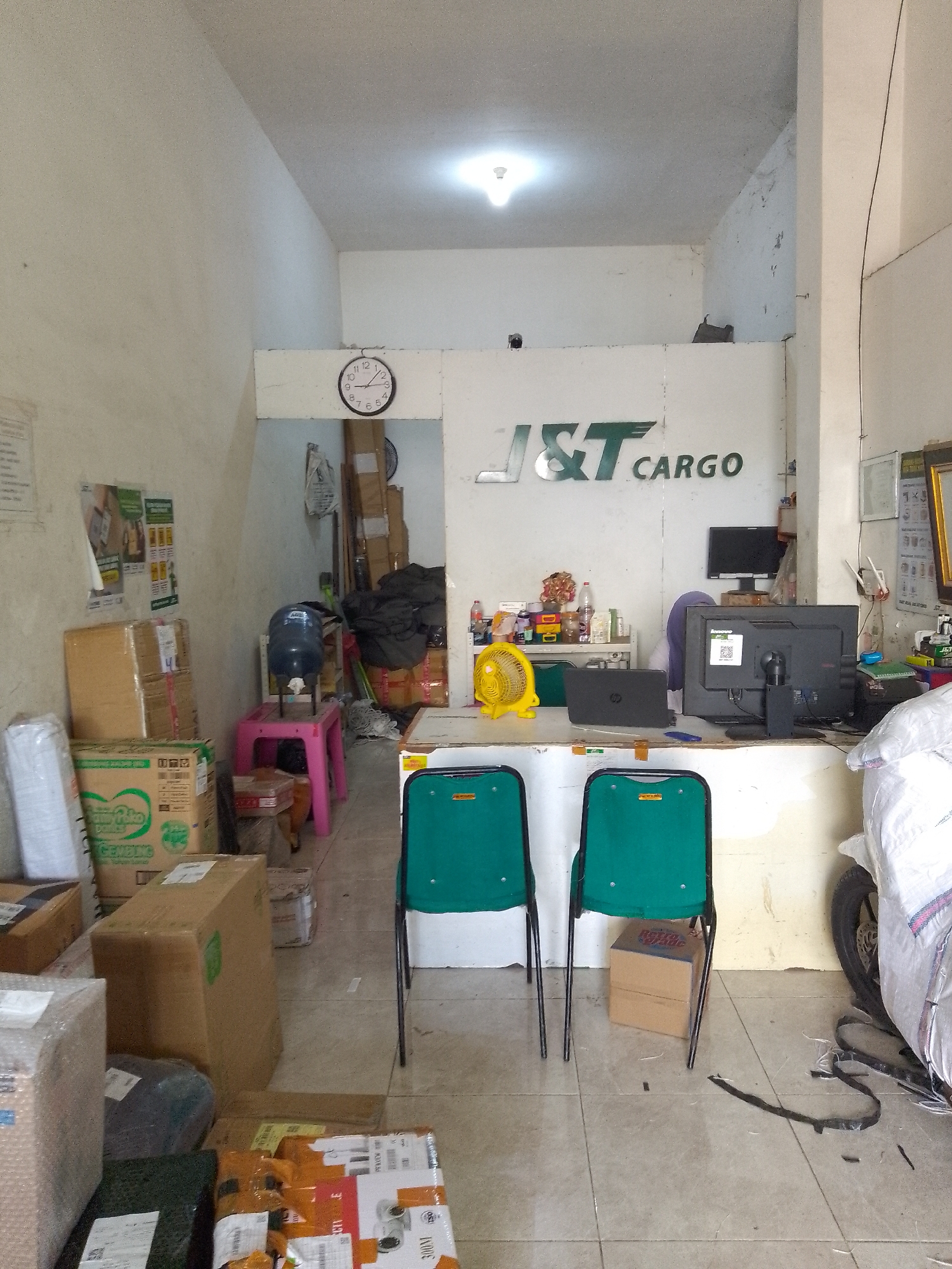 The image depicts the interior of a logistics company, likely a J&T Cargo branch, located in Tumenggungan, Indonesia. The scene is characterized by a cluttered yet functional workspace. In the foreground, two empty green chairs with black metal frames face a counter. A brown cardboard box is placed beneath the chair on the right. The floor is tiled.
Behind the chairs is a counter area. On the counter are two computer monitors, a black laptop, a yellow fan, and various items including bottles and what appear to be office supplies. A partially visible person wearing a purple headscarf is seated behind the counter, tending to the computer.
The back wall prominently features the "J&T CARGO" logo in green lettering. To the left of the counter, several stacked cardboard boxes and other packages are visible. A water dispenser with a large blue bottle sits on a pink plastic stool. Further to the left, posters are affixed to the wall, and more packages are stacked.
In the background, additional stacked boxes and bags are present, suggesting a storage or processing area. A wall clock is mounted above the counter area. To the far right, a covered motorcycle is parked, its wheels and parts of the cover visible. The overall environment appears to be an indoor commercial space with limited natural light, relying on overhead artificial lighting. The scene suggests a busy operational environment for package handling and delivery services.