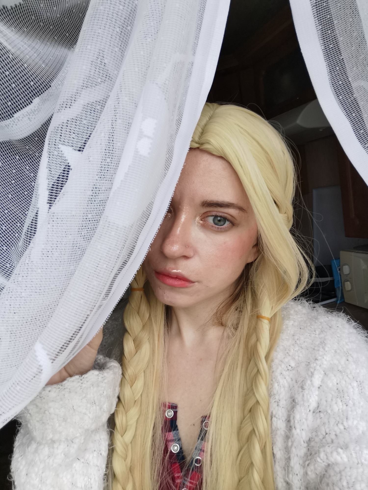 A woman with blonde braided hair is partially obscured by white netting. She is looking at the camera with blue eyes and is wearing a white fuzzy sweater over a plaid shirt. Her makeup includes pink blush and lip gloss. The background appears to be indoors, possibly a kitchen, with cabinets and an appliance visible. The netting is draped in front of her, creating a soft focus effect.