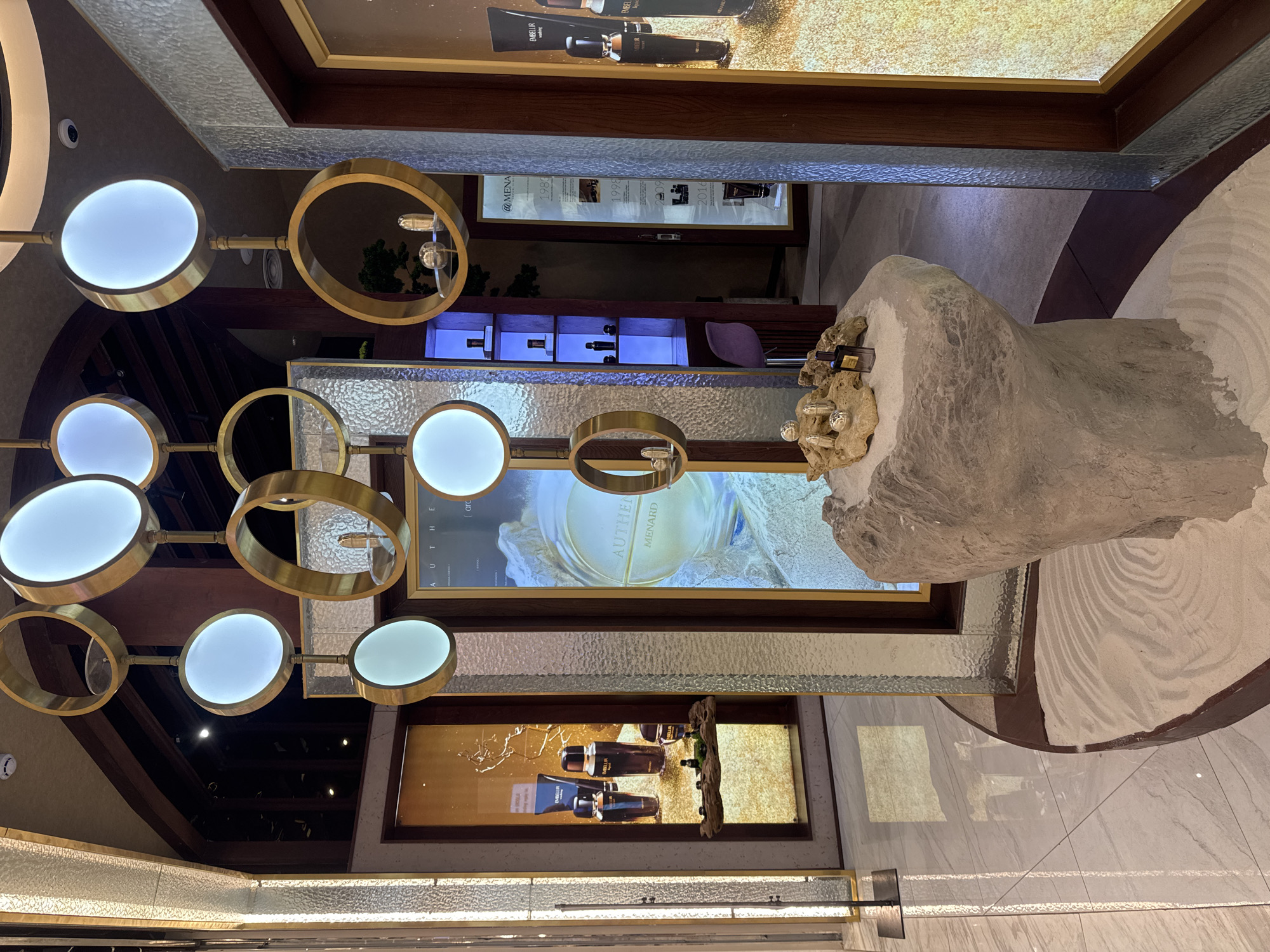 The image depicts the interior of a luxury cosmetics store, likely a Menard boutique, in Hanoi, Vietnam. The store features a minimalist and elegant design with a focus on natural elements and soft lighting. A prominent feature is a large, light-colored stone sculpture serving as a display pedestal, adorned with several small cosmetic bottles and a perfume bottle. Above this, a modern chandelier with circular gold frames and illuminated white discs hangs, casting a warm glow.
The background reveals display cases and promotional screens showcasing Menard skincare products. One screen prominently displays a circular logo with the text "AUTHENTIC MENARD". Another display to the left shows several bottles and tubes of Menard cosmetics against a textured gold background. In the far background, a doorway leads to another section of the store, where shelves with more products are illuminated by blue light. The flooring is a combination of polished white tiles and dark wood accents, with a section of raked white sand in the foreground, adding to the tranquil aesthetic. The overall impression is one of sophistication and a serene shopping experience.
