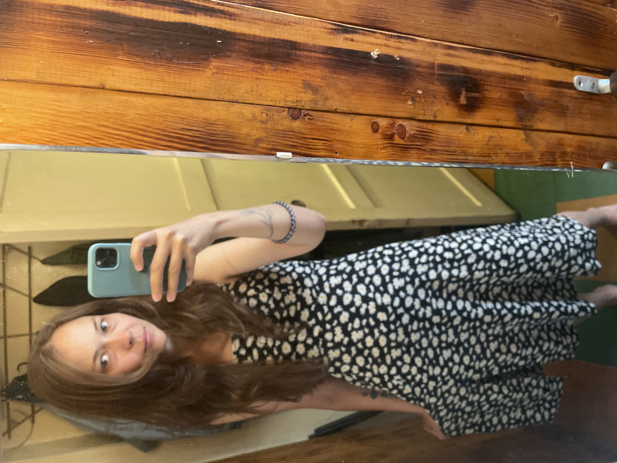 A woman with long, wavy brown hair stands in front of a mirror, taking a selfie with a light blue smartphone. She is wearing a black dress with a white daisy pattern and appears to be in a room with wooden walls and a door. The reflection shows a coat rack in the background.