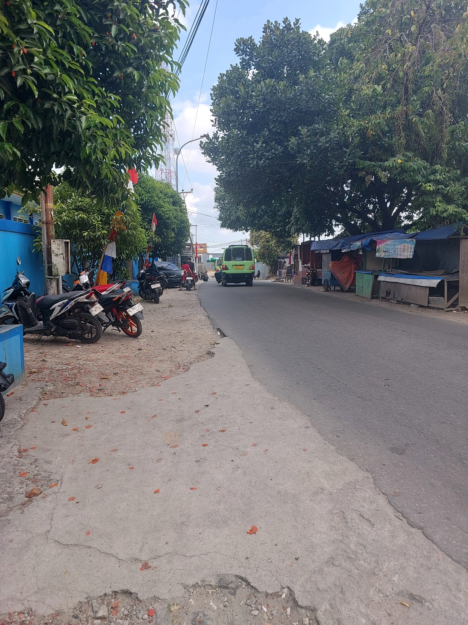 The image shows a street scene in Ambon, Indonesia. In the foreground, several motorcycles are parked on a cracked and sandy sidewalk, some with red and white Indonesian flags attached. A bright blue building is visible on the left, with a portion of a telecommunications tower in the background. The street itself is paved and appears to be a two-lane road with a slight incline. On the right side of the street, there are several small market stalls with tarpaulin roofs selling various goods. A green van is driving away from the camera on the right side of the road, and a person is riding a red motorcycle in the distance. Large trees with dense green foliage line the street, and the sky is a clear blue with a few scattered clouds.