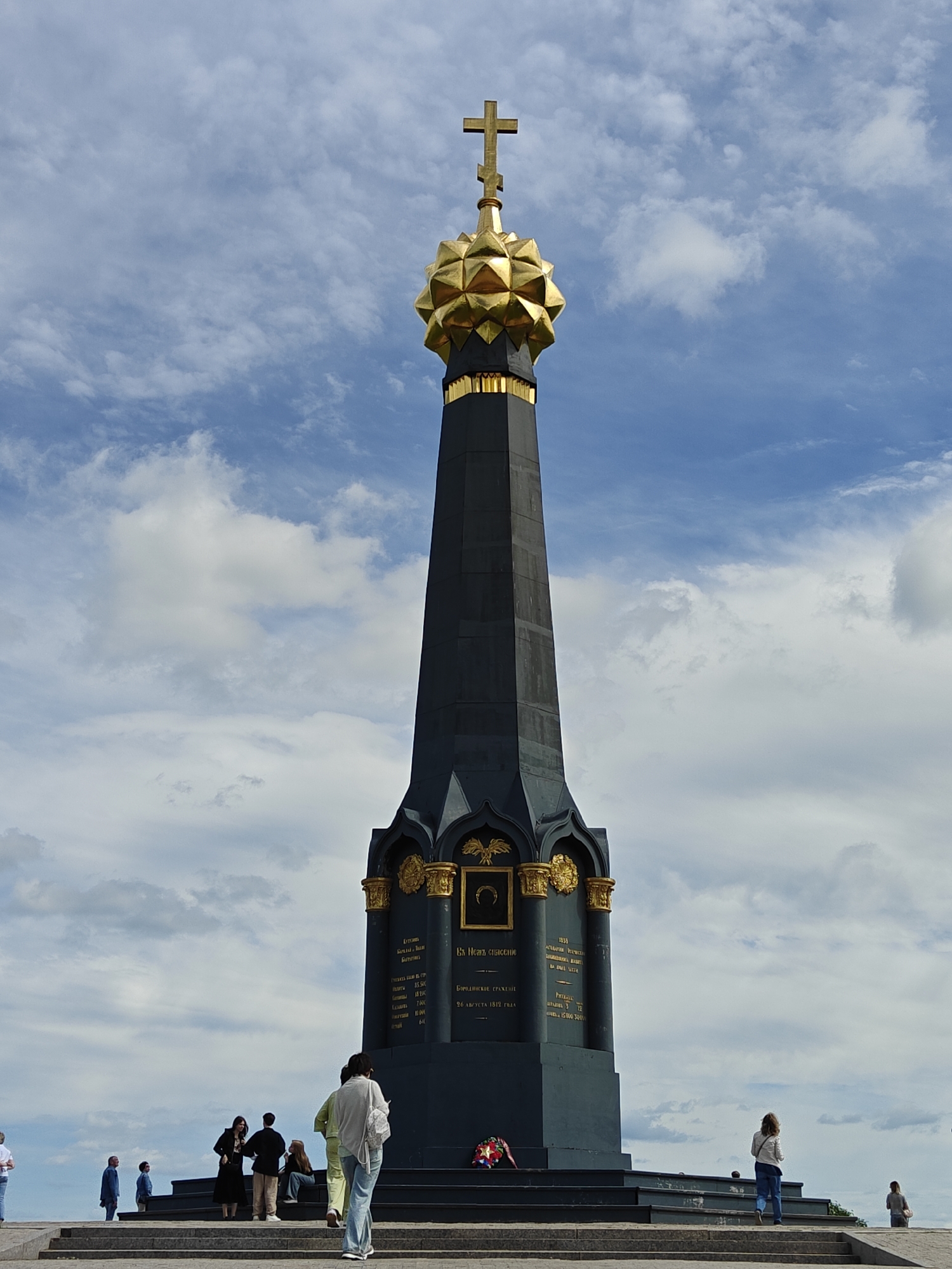 The image shows the Main Monument at the Borodino Battlefield in Borodino, Russia. It's a tall, black column-like structure with golden ornamental details, topped with a golden orb and a cross. The monument is surrounded by a low, tiered platform with people standing around its base. Some people are looking at the monument. The sky is blue with scattered white clouds.