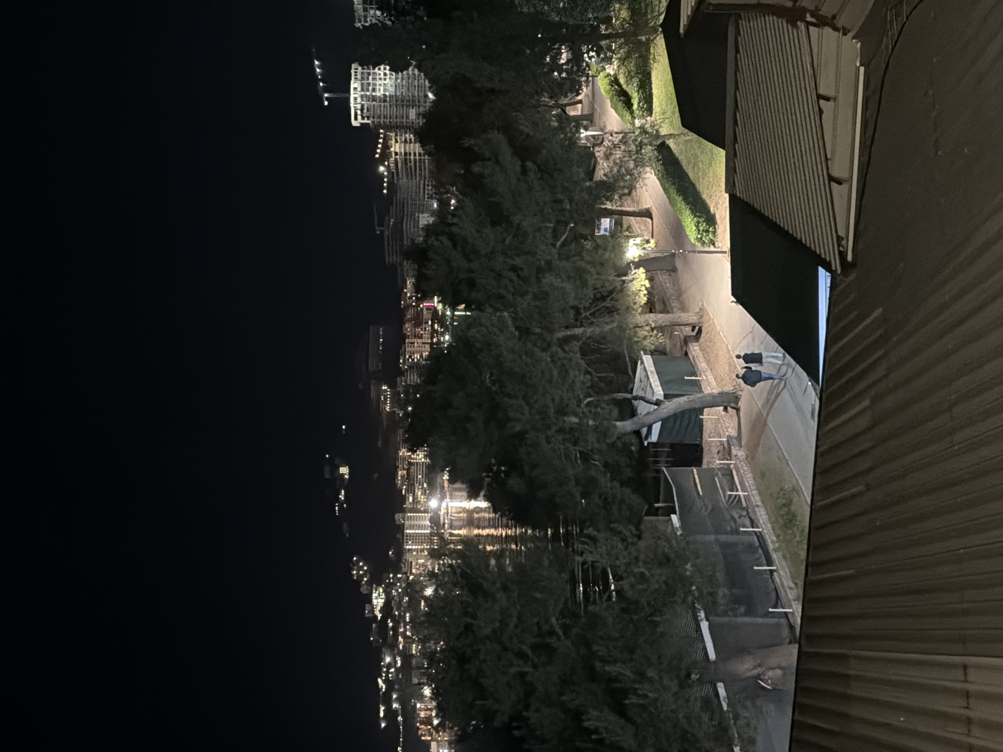 The image presents an elevated nighttime view from a structure, potentially a building, in Budva, Montenegro. In the distance, the illuminated cityscape of Budva reflects on the water. Trees obscure portions of the view between the building and the cityscape. Closer to the vantage point, a path is illuminated by streetlights. Two people are seen walking along the path. The foreground includes the corrugated metal of the building's structure.