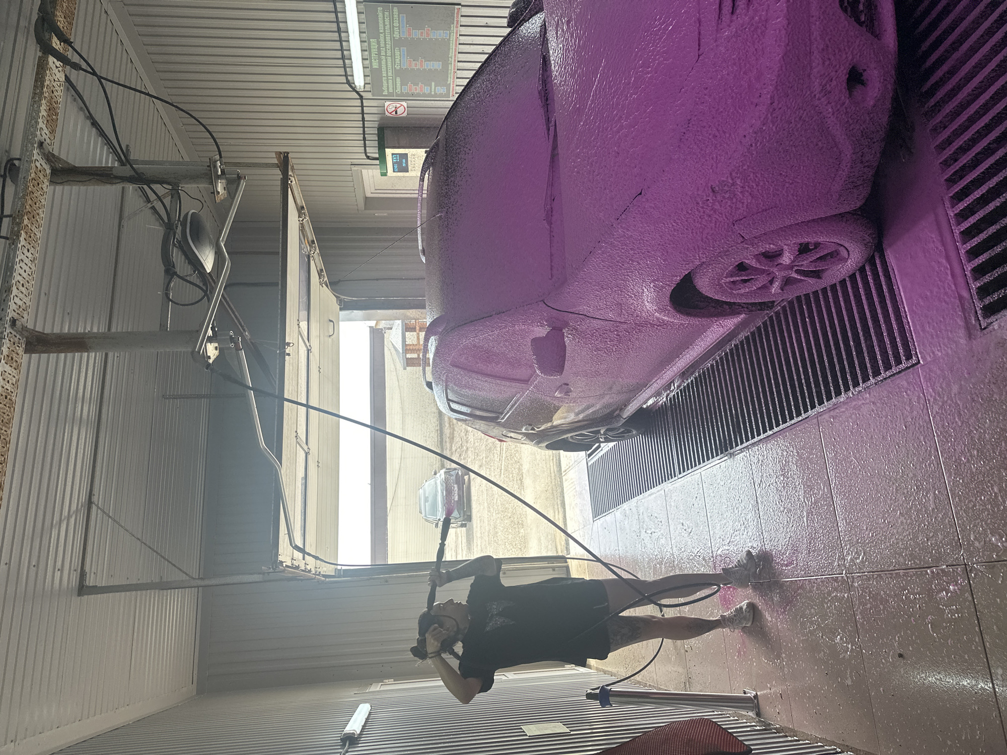 The image shows a car wash facility, likely located in Chaikovsky, Russia, given the presence of Russian text on a sign. A car covered in purple foam occupies a large portion of the frame, positioned on a grate for drainage. A person is present, holding a high-pressure washer and aiming it at the ground, presumably cleaning the area. The facility features corrugated metal walls and ceiling. There are overhead structures and equipment associated with the car wash process. Outside the car wash bay, a street scene is visible with another car parked. The setting appears to be an enclosed structure designed specifically for vehicle cleaning.