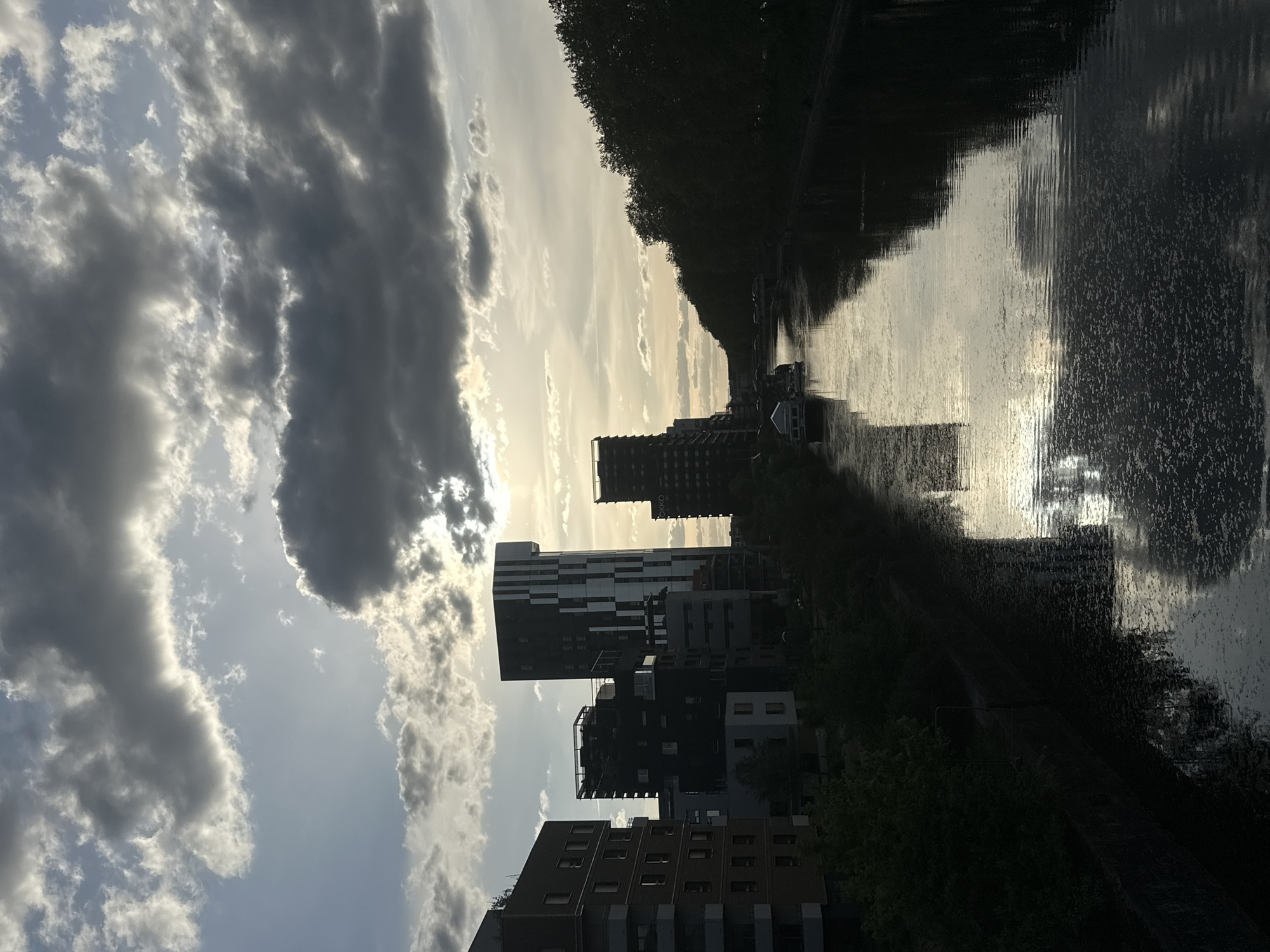 The image shows a canal scene in Strasbourg, France. The canal reflects the sky and some buildings. Modern apartment buildings line one side of the canal. Trees partially obscure the far side of the canal, but some boats are visible. The sky is filled with clouds, and the sun is bright behind them.