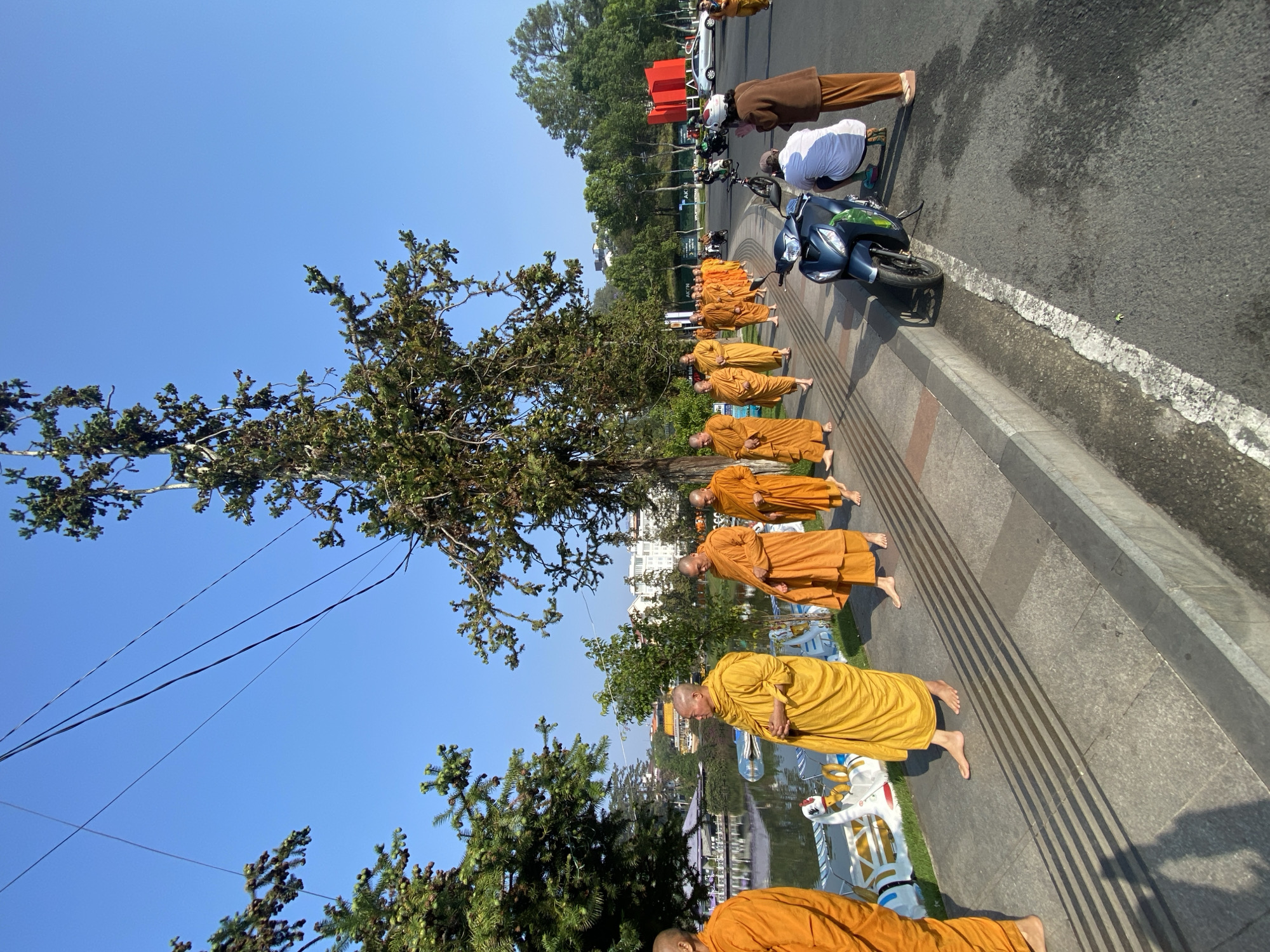 A line of monks in saffron robes are kneeling on a paved street next to a lake. A blue motorbike is parked on the road. In the background, buildings and trees are visible. The location is Da Lat, Vietnam.
