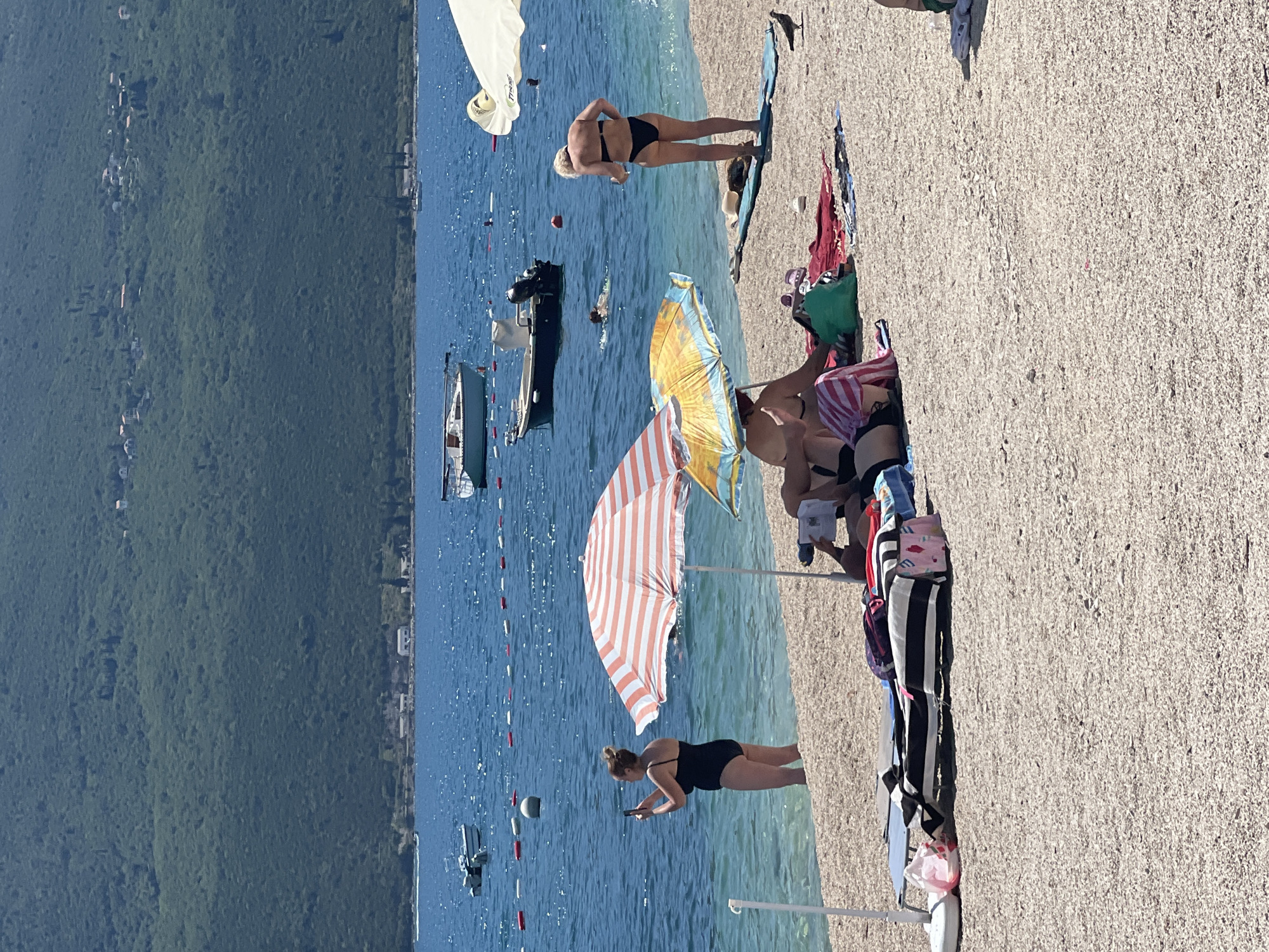 The image depicts a sunny day at a pebbled beach in Baošići, Montenegro. The Adriatic Sea, a deep blue, stretches to the horizon, where a green, tree-covered mountain range forms the backdrop. Several small boats are anchored or moving on the water, and a line of buoys marks a boundary in the sea.
On the beach, several people are enjoying the sun. In the foreground, a woman in a black swimsuit stands at the water's edge, holding a phone to take a picture. Two beach umbrellas, one striped orange and white and the other yellow and orange, provide shade. Under the orange and white umbrella, several people are partially visible, with towels and bags laid out. To the right, a woman in a black bikini stands on the sand. In the middle ground, more people are relaxing on towels, some under umbrellas. A pigeon is visible on the sand to the right of the woman in the black bikini. The ground is covered in small white and grey pebbles.