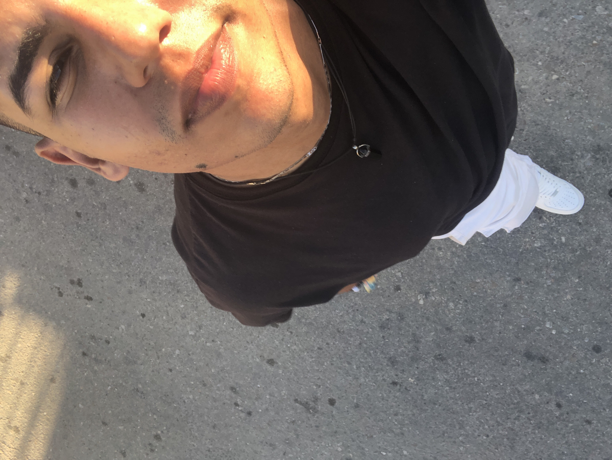 A high-angle shot depicts a person standing outdoors on what appears to be asphalt or concrete pavement. The individual, facing upwards towards the camera, is partially visible from the face down to the mid-torso and partially includes one foot. They are wearing a dark-colored, possibly black, t-shirt, and white shorts or pants. White sneakers are also visible on the foot. The person has fair skin, dark hair, and is wearing a thin necklace. The background is the textured surface of the ground with some minor spots or discoloration. The image has strong directional lighting. There is no specific evidence to confirm the location is Altagracia, Venezuela.